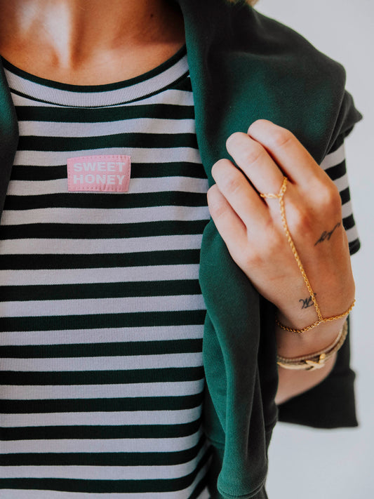 Model is wearing a green and lavender striped two-piece set with a green sweater draped over her shoulder. Up close picture of the SweetHoney logo tag in pink and white in the center of chest.