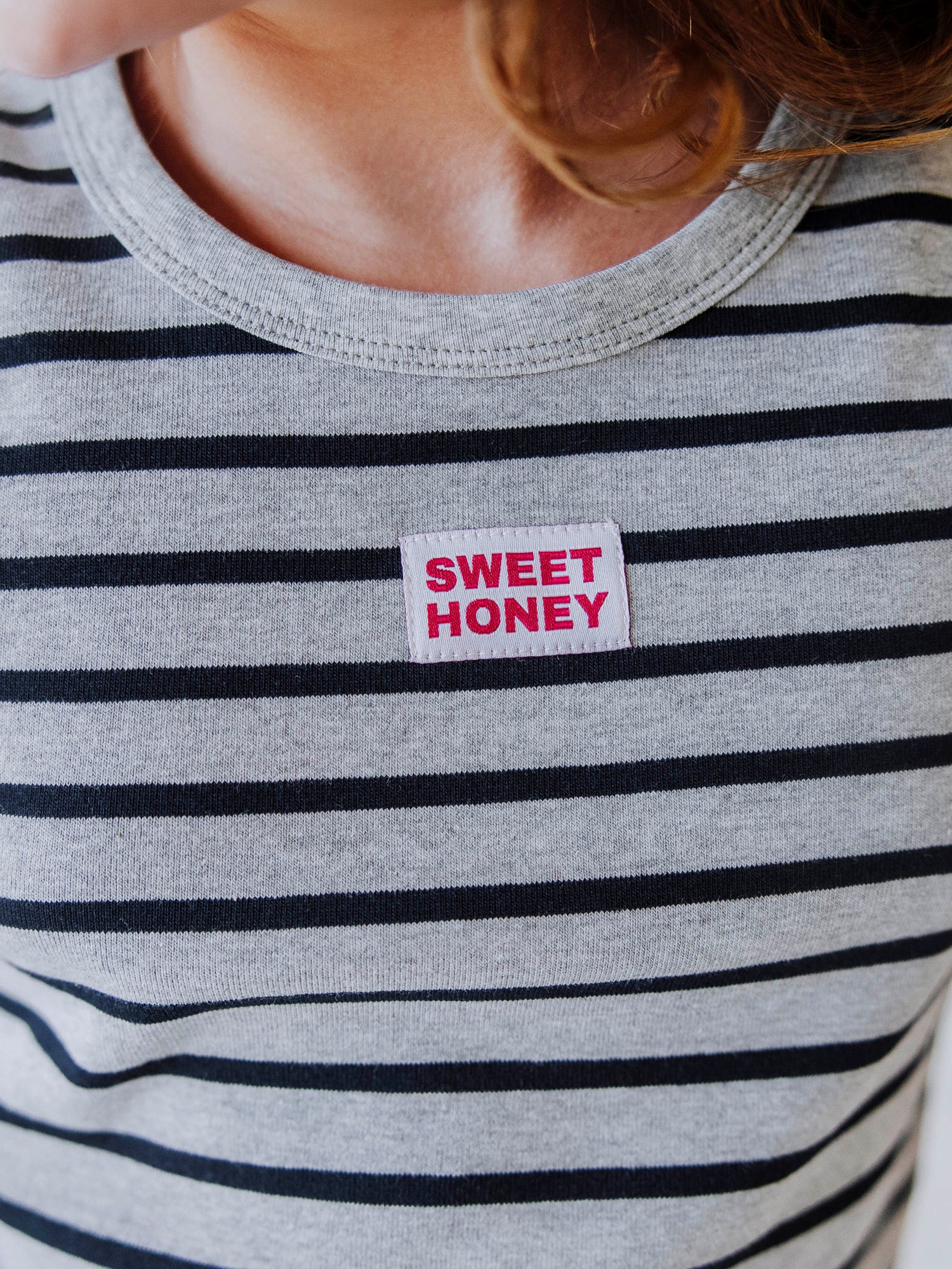 Up close picture of a gray and navy striped shirt and centered in the middle is a sweethoney tag that is red and white. 