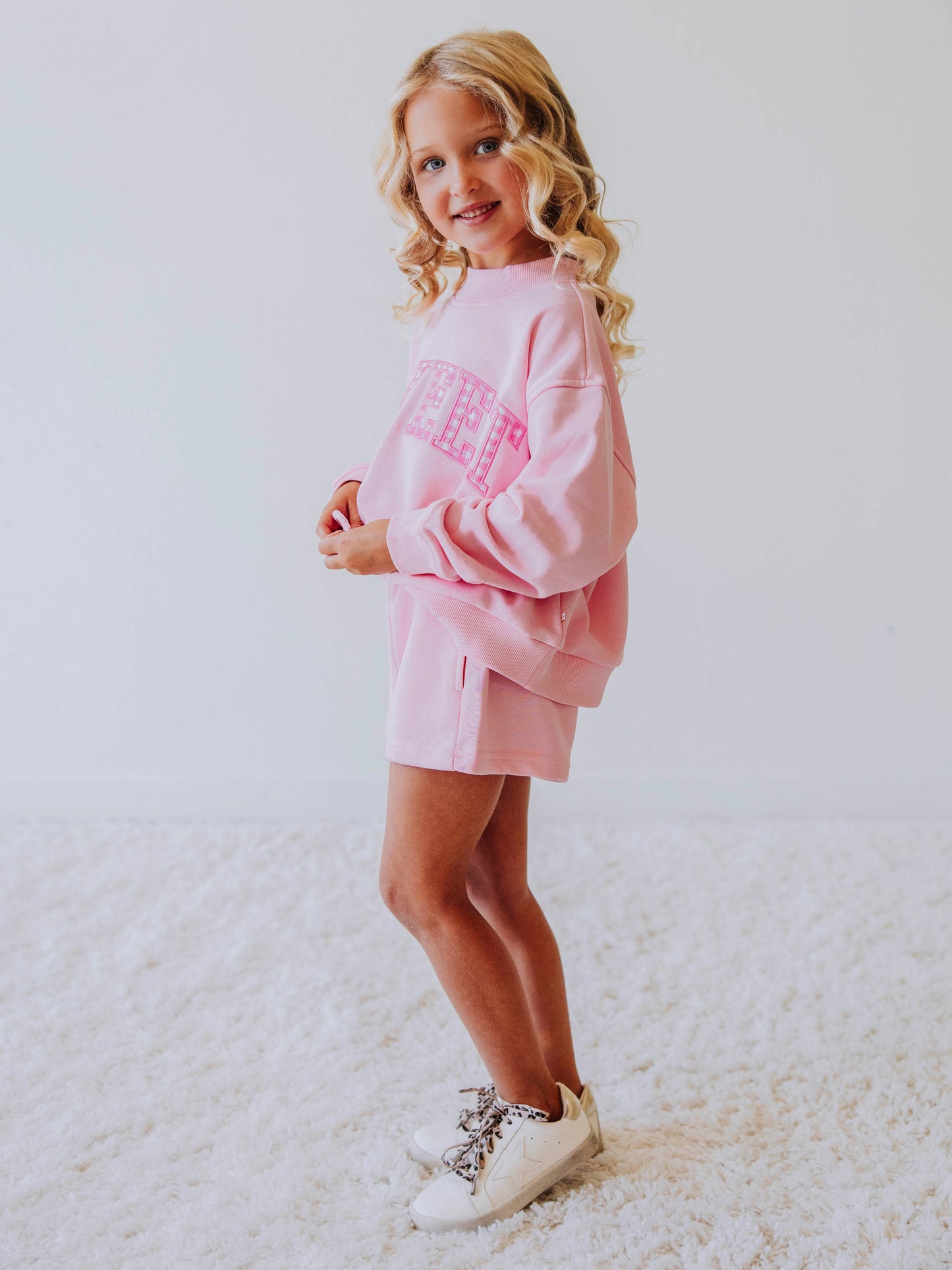 Girl is wearing a two-piece pink jogging set. The top says SWEET on the front and the letters have pink gingham checks in each. Bottoms are the same pink as top. Side angle of model. 