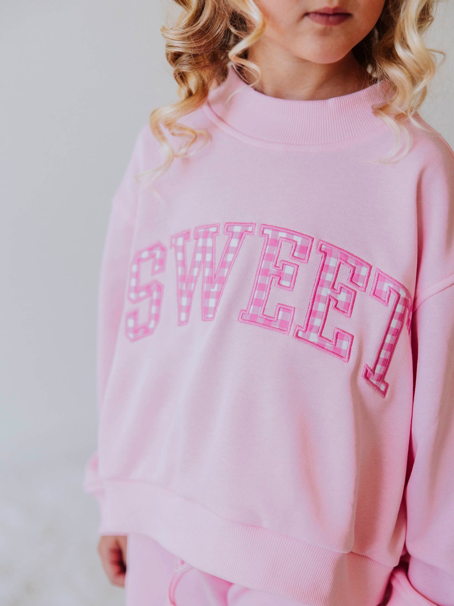 Close up of model wearing a pink mock neck sweatshirt that says SWEET on the front. In each letter are pink gingham checks. 