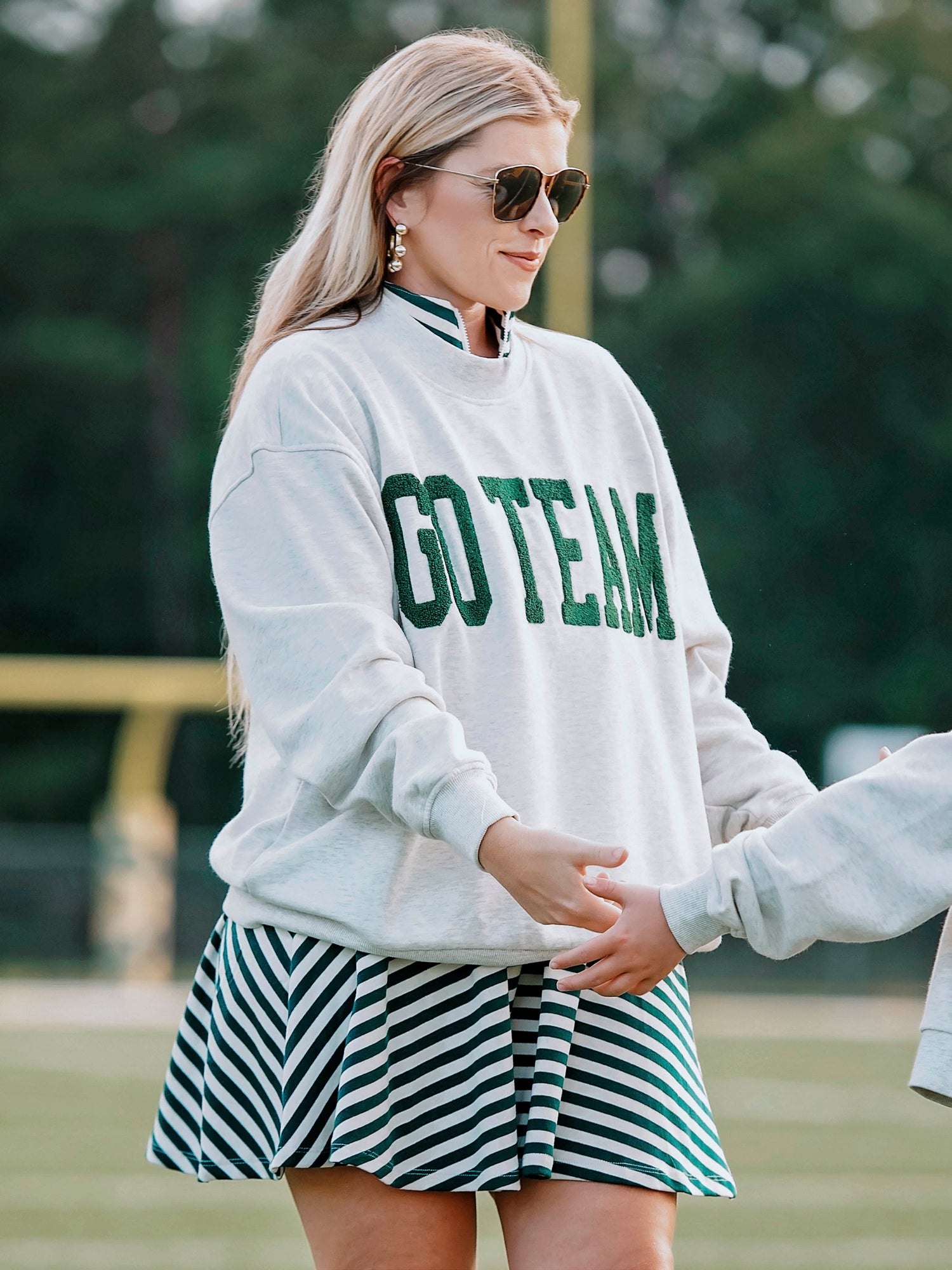 Women outside wearing a heathered gray sweatshirt that says GO TEAM in dark green. She is wearing the sweater over a green and white striped dress