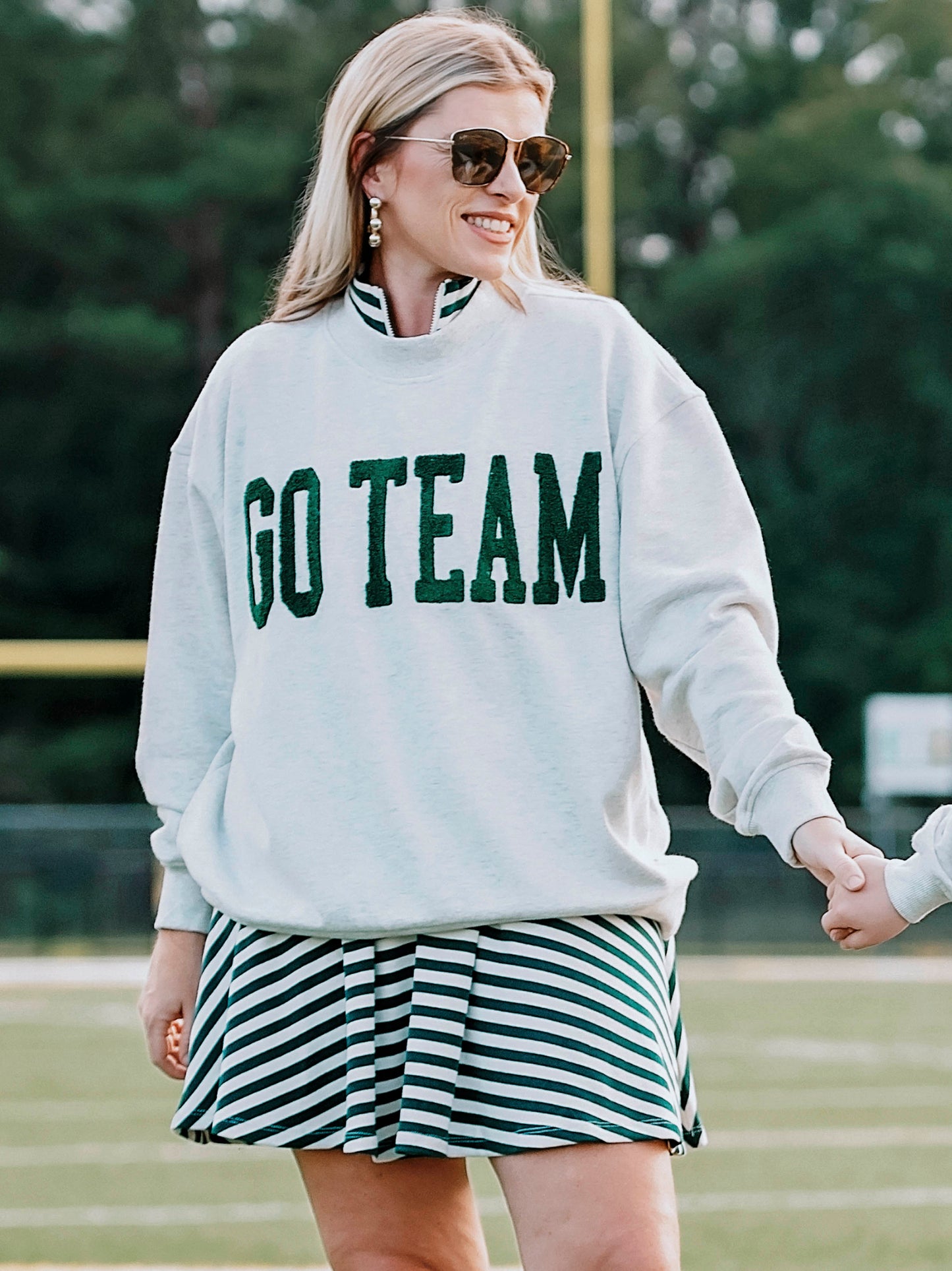 Women outside wearing a heathered gray sweatshirt that says GO TEAM in dark green. She is wearing the sweater over a green and white striped dress