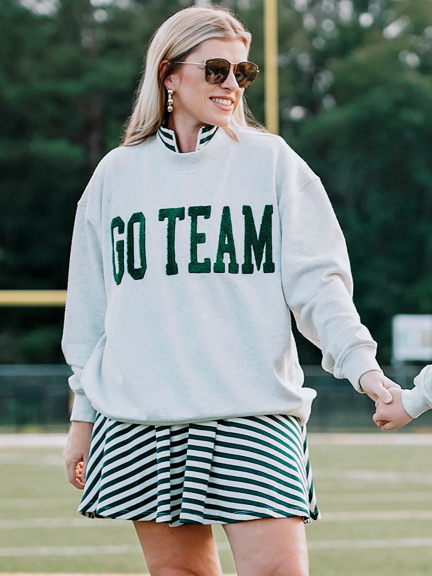 Women outside wearing a heathered gray sweatshirt that says GO TEAM in dark green. She is wearing the sweater over a green and white striped dress