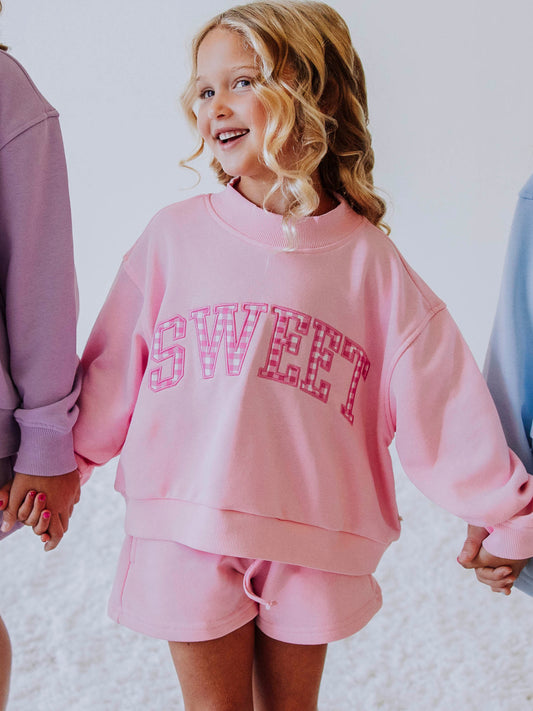 Girl is holding hands with two others and is wearing a two-piece, pink set. Top says SWEET with gingham checks in each letter. Bottoms are the same color as the top. 