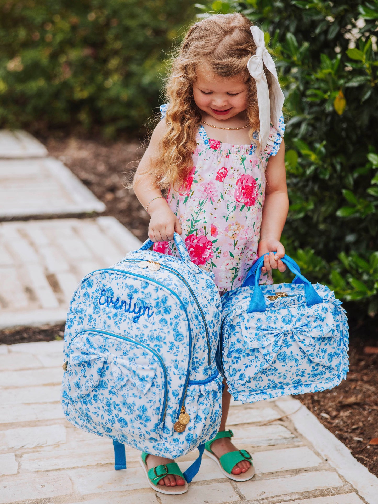 Girl standing outside holding both a blue floral backpack and lunch kit. Both have an attached bow on fronts.