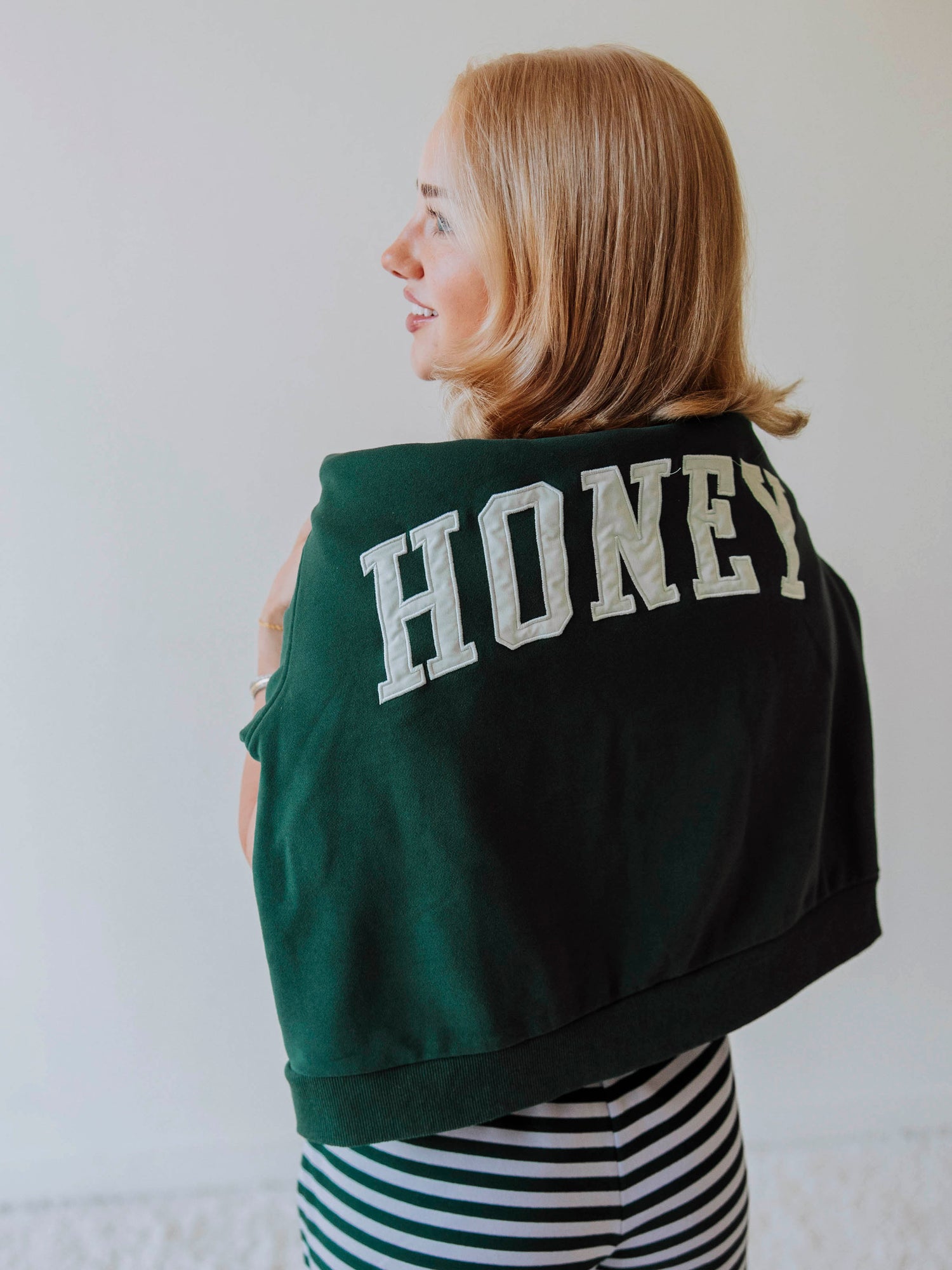 Girl showing a green mock neck sweatshirt  that says HONEY around her shoulders. Bottoms appear to be striped.