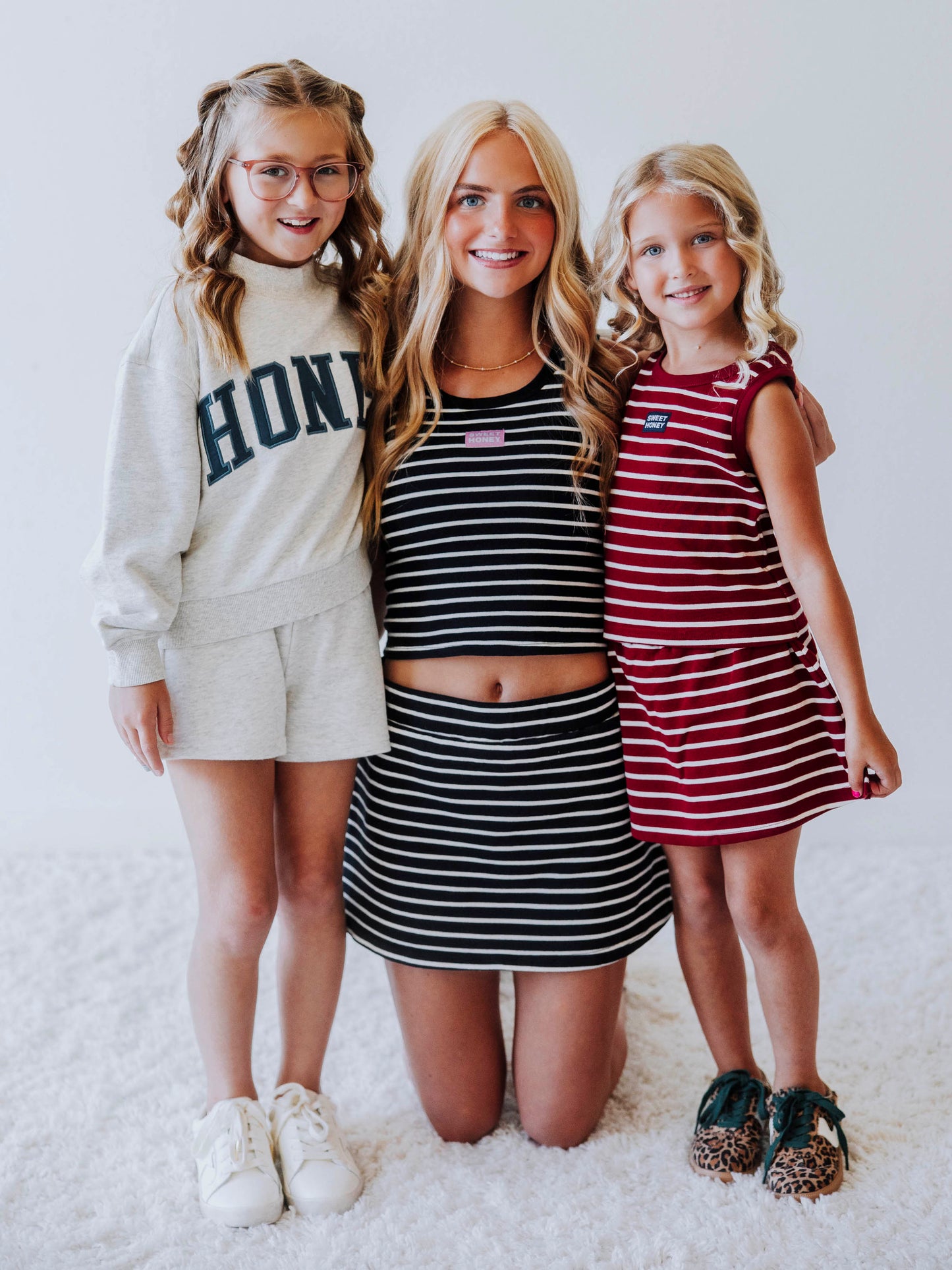 Pictured are three models. Model on left is wearing a mock neck sweatshirt that says honey on the front in green. Shorts she is wearing is the same heathered gray. Model in middle is wearing a navy and ivory striped skort set with a stittched SweetHoney logo tag on center of chest. Model on right is wearing a crimson and ivory striped skort set too with stitched sweethoney tag on chest