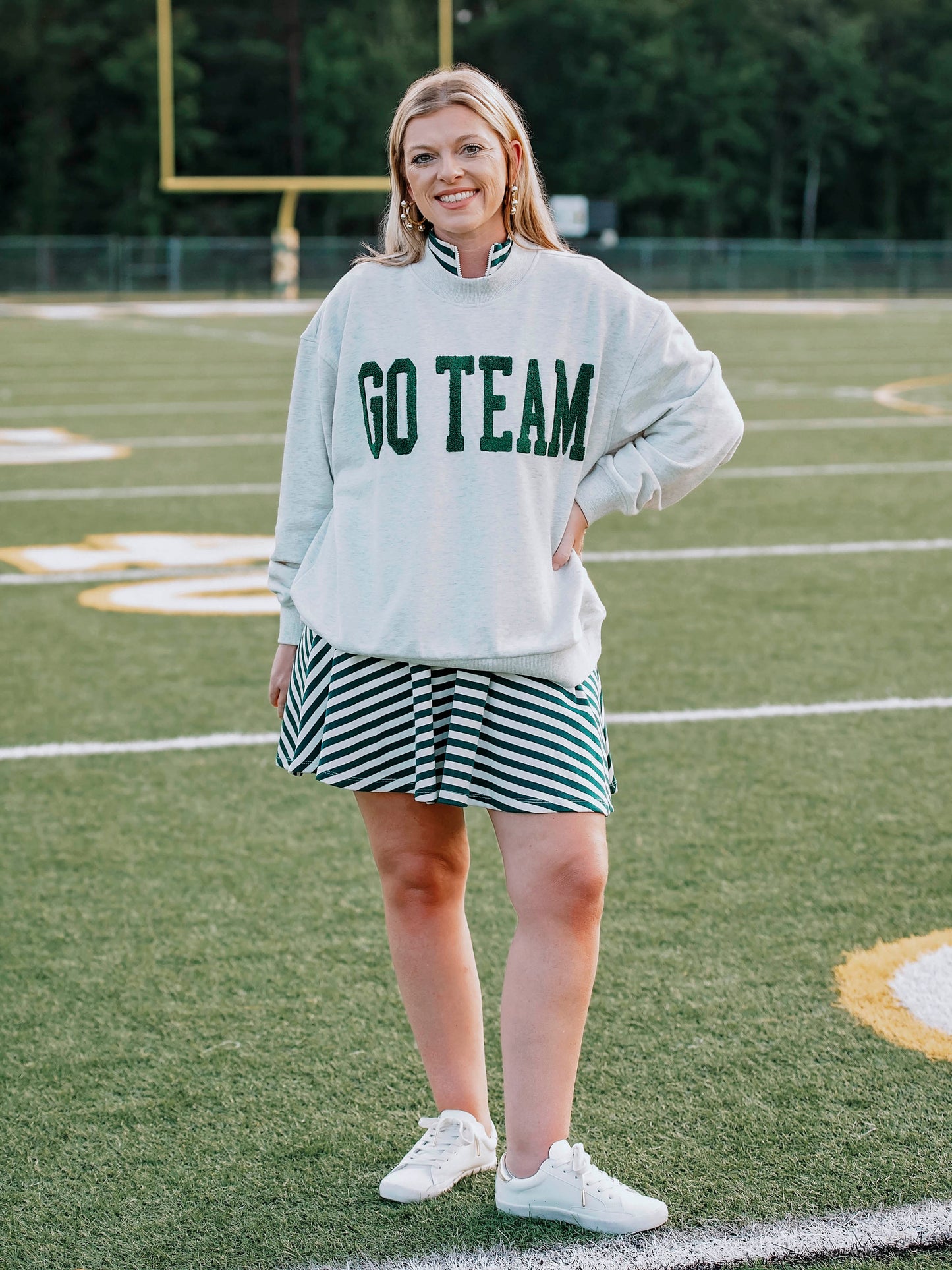 Women outside wearing a heathered gray sweatshirt that says GO TEAM in dark green. She is wearing the sweater over a green and white striped dress
