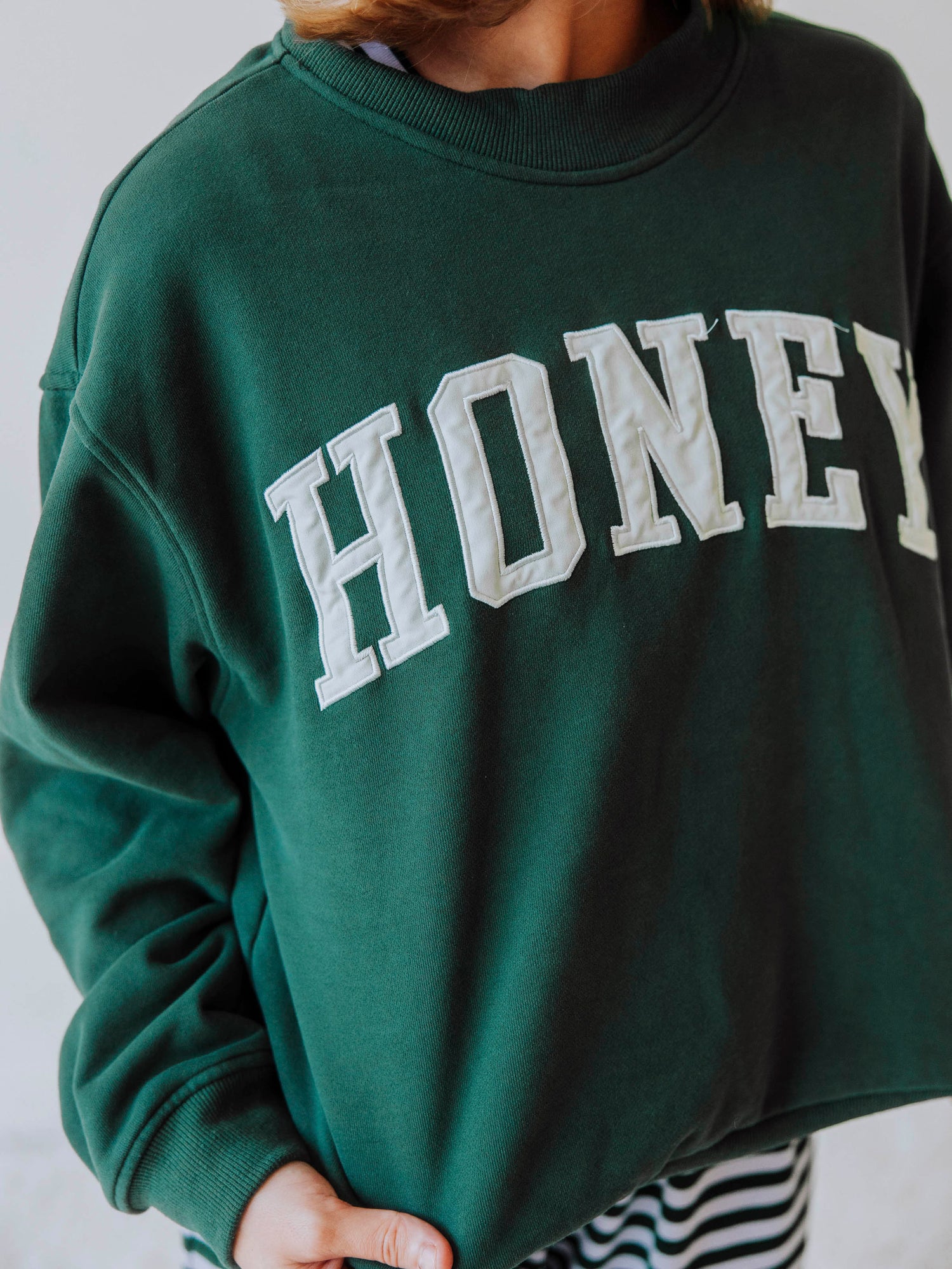 Girl wearing a green mock neck sweatshirt  that says HONEY on the front. Bottoms appear to be striped.