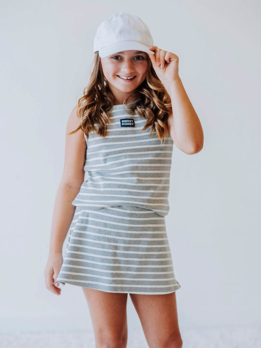 Model is wearing a two-piece gray and white striped skort set. There is a SweetHoney tag that is centered on front of shirt and is navy and white in color. Model is also wearing a white hat. 