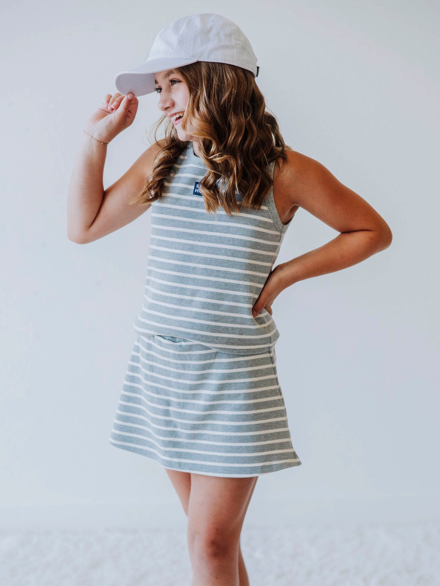 Model is wearing a two-piece gray and white striped skort set. There is a SweetHoney tag that is centered on front of shirt and is navy and white in color. Model is also wearing a white hat. 