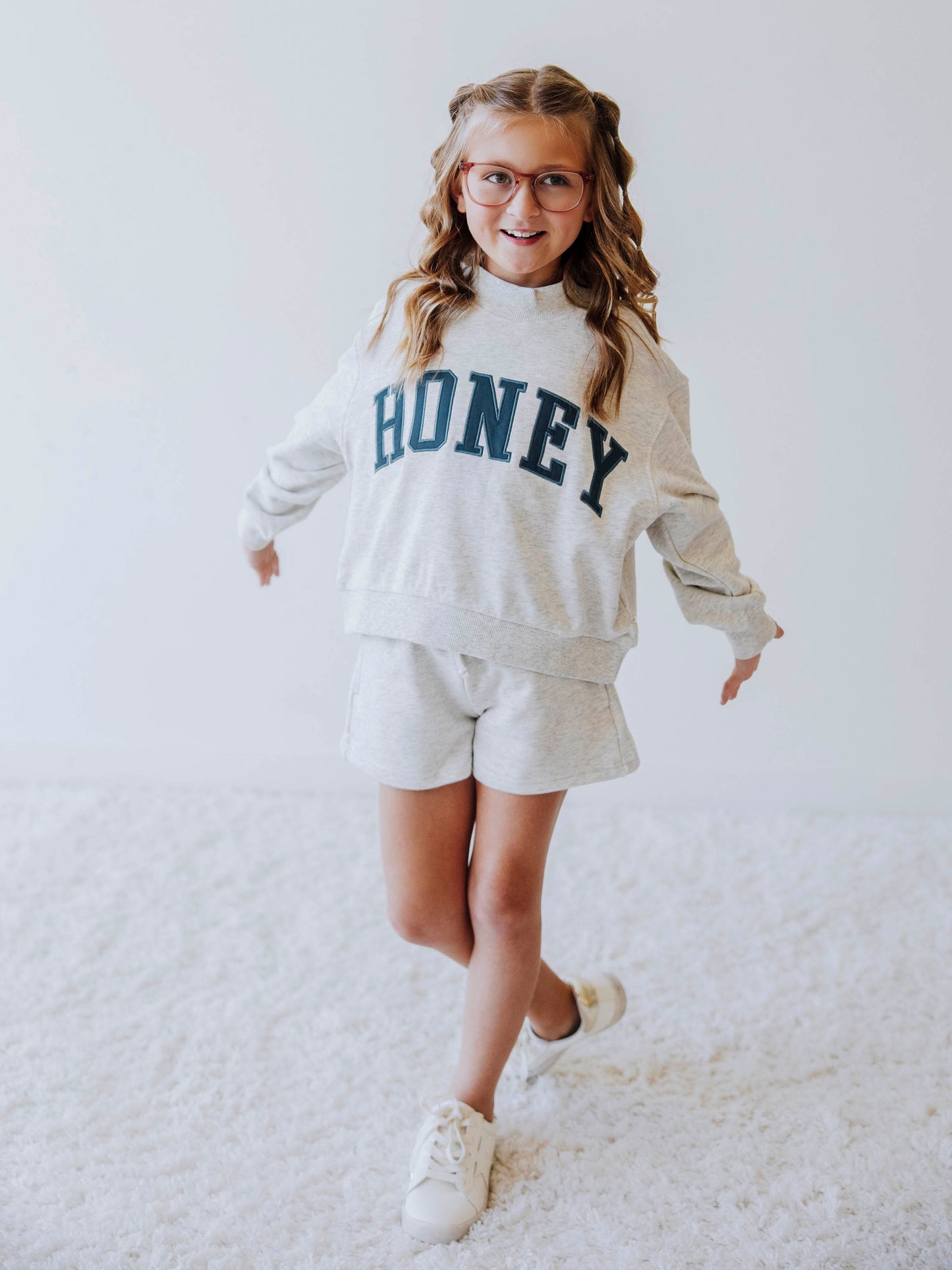 Girl model is wearing a two piece, gray, mock neck style sweat set. The top says HONEY across the top in a greenish color. Model is also wearing white shoes. 