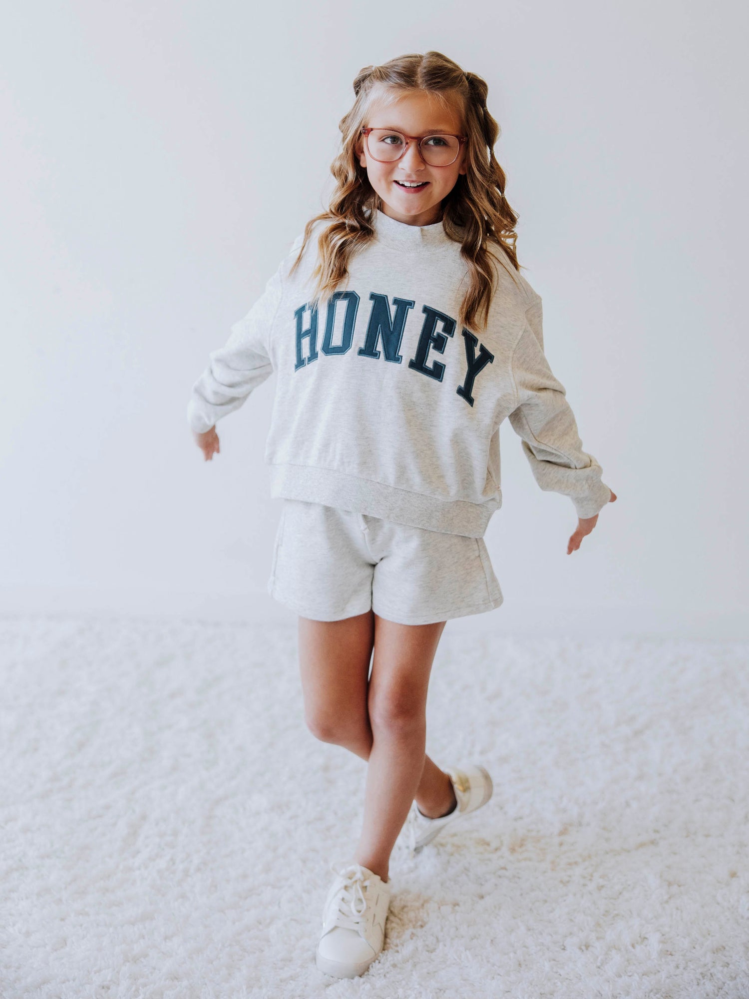 Girl model is wearing a two piece, gray, mock neck style sweat set. The top says HONEY across the top in a greenish color. Model is also wearing white shoes. 