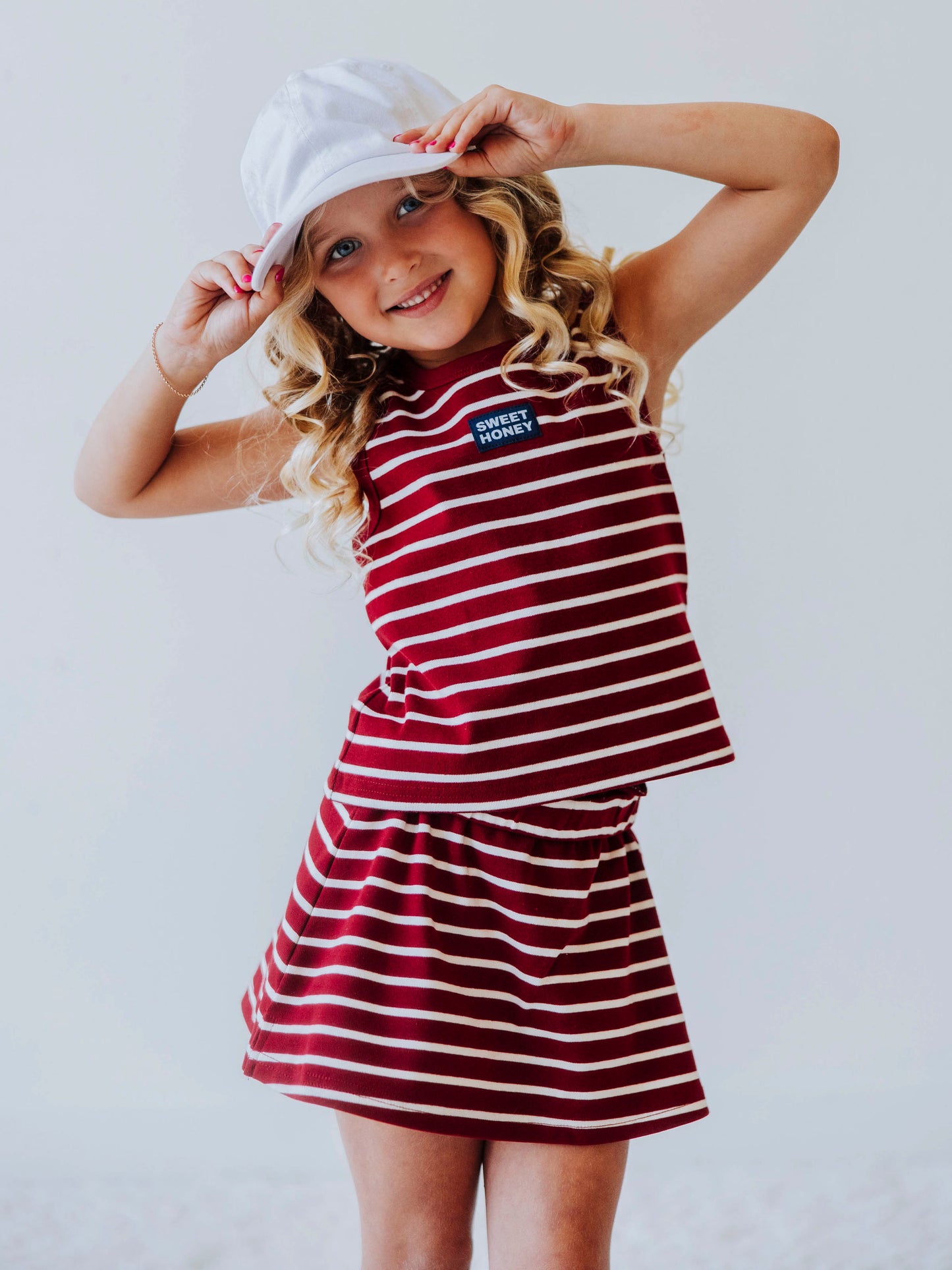 Model is wearing a two-piece, crimson and ivory striped skort set. Model is wearing a white hat. Centered on the shirt is a SweetHoney tag that is navy and white in color. 