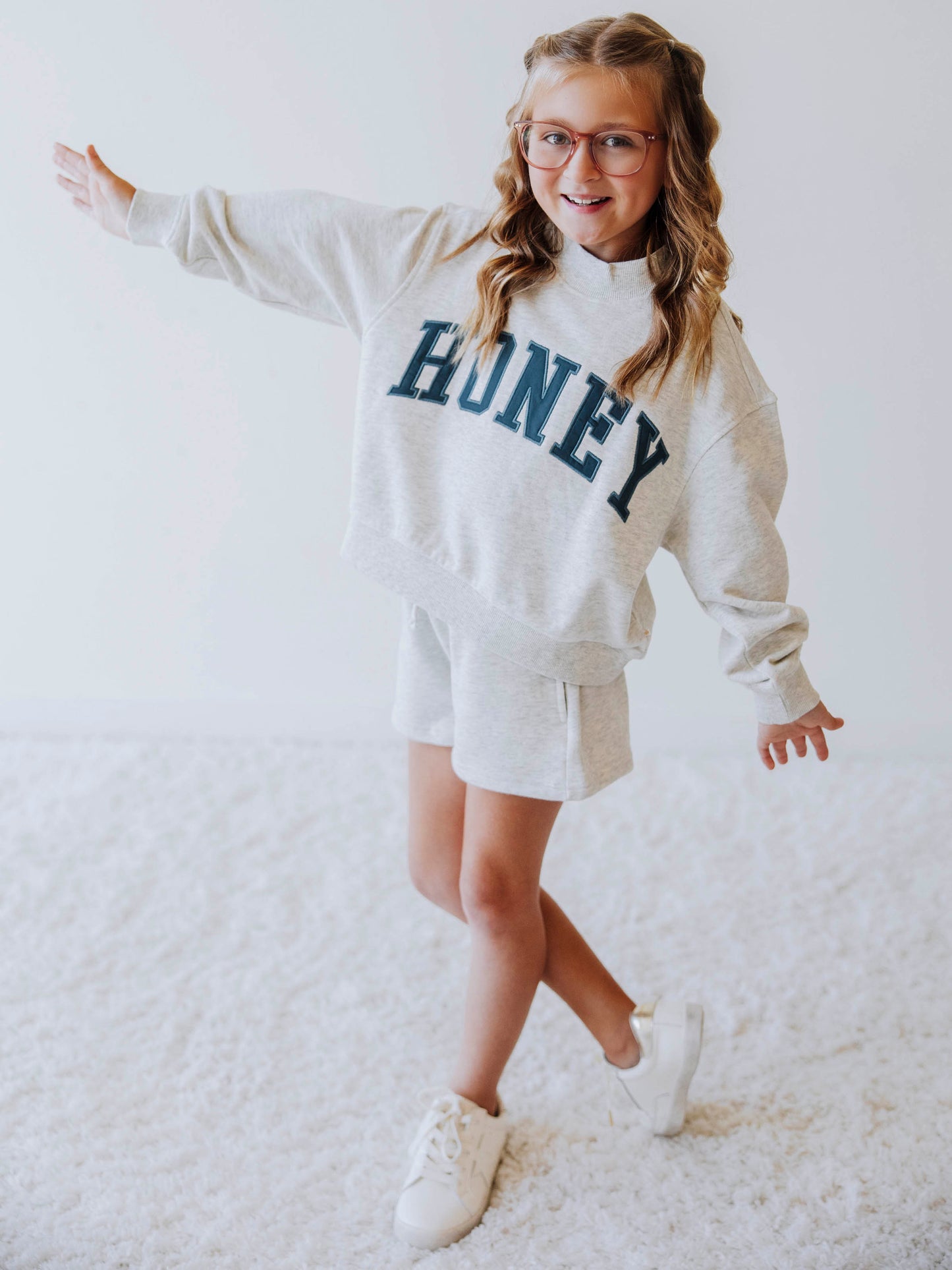 Girl model is wearing a two piece, gray, mock neck style sweat set. The top says HONEY across the top in a greenish color. Model is also wearing white shoes. 