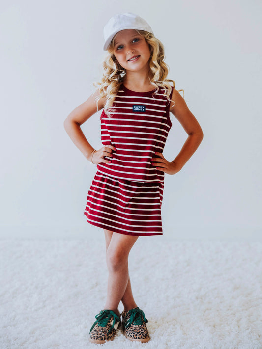 Model is wearing a two-piece, crimson and ivory striped skort set. Model is wearing a white hat. Centered on the shirt is a SweetHoney tag that is navy and white in color. Model has on leopard shoes with green laces. 