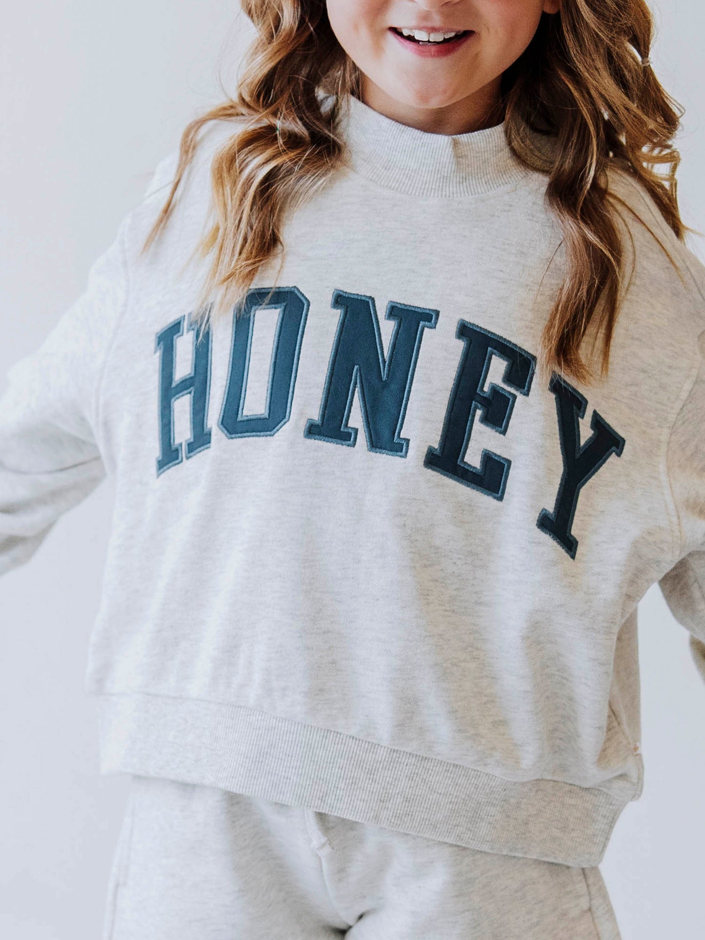 Close up of Girl model wearing a grey mock neck style shirt that says HONEY across the top in a dark greenish color. 