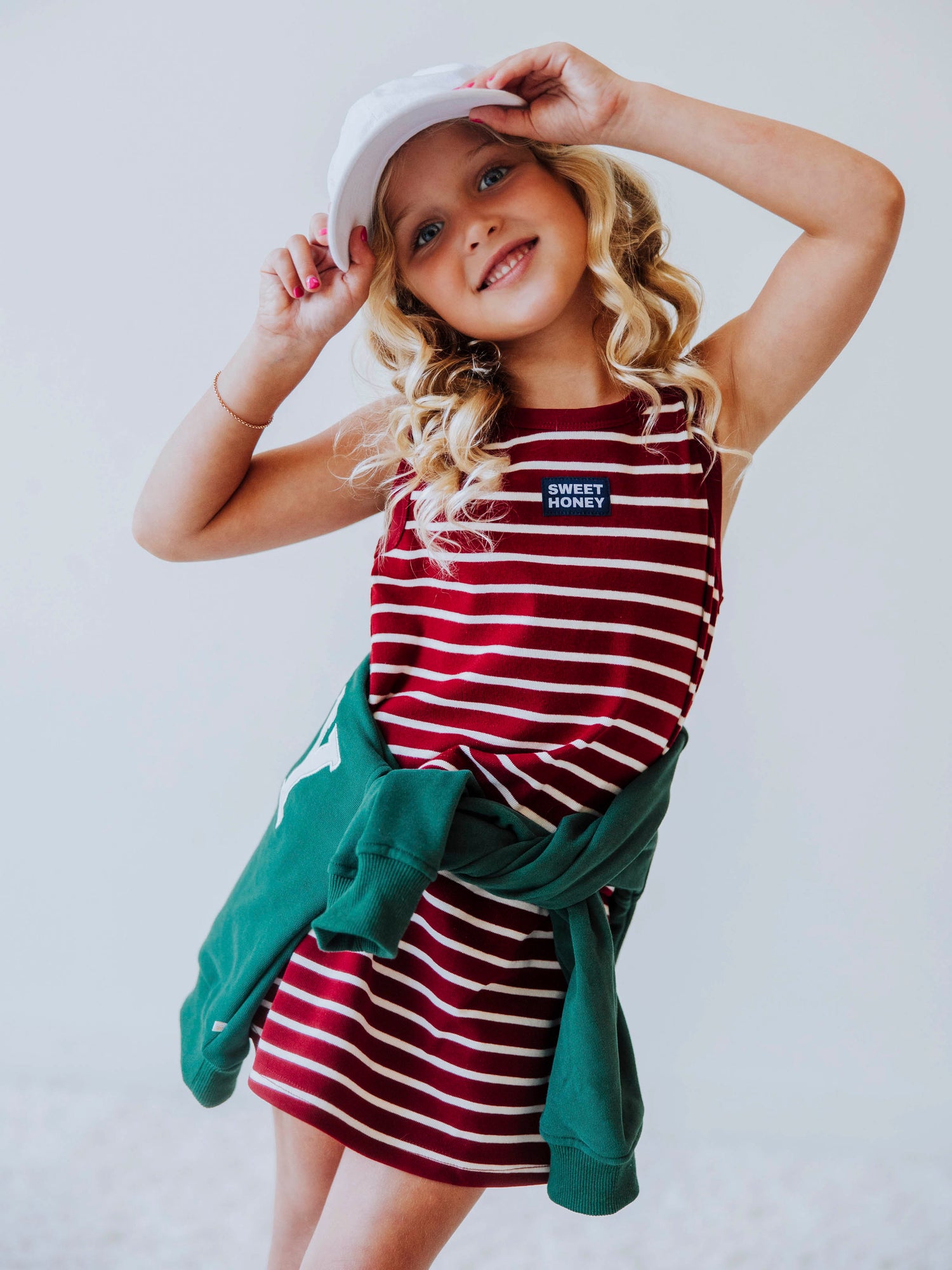 Model is wearing a two-piece, crimson and ivory striped skort set. Model is wearing a white hat. Centered on the shirt is a SweetHoney tag that is navy and white in color. Green sweater around waist. 