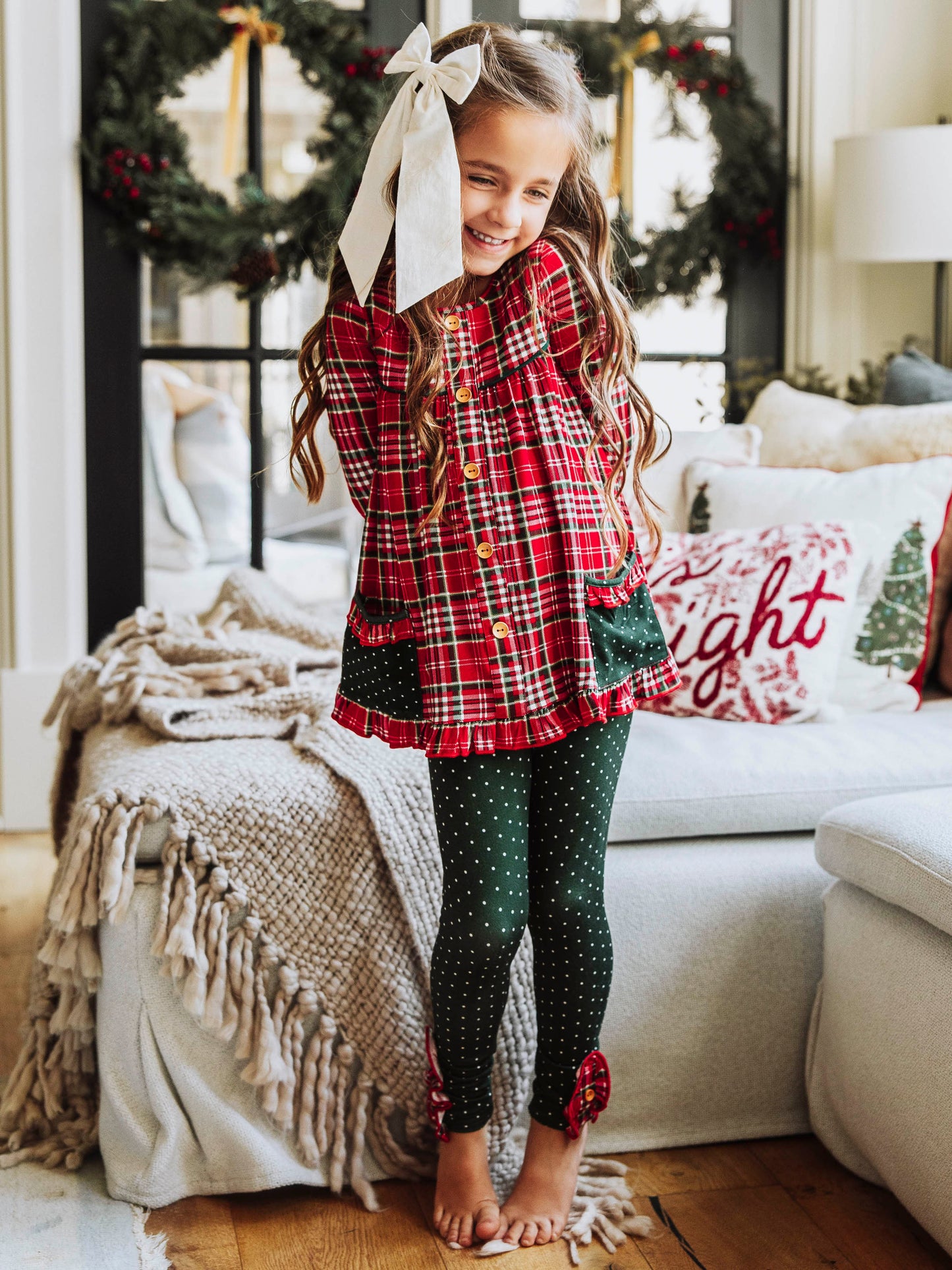Girl wearing a two piece, Christmas play set. Top is a traditional plaid Christmas print and the bottoms are dark green polka dot. 
