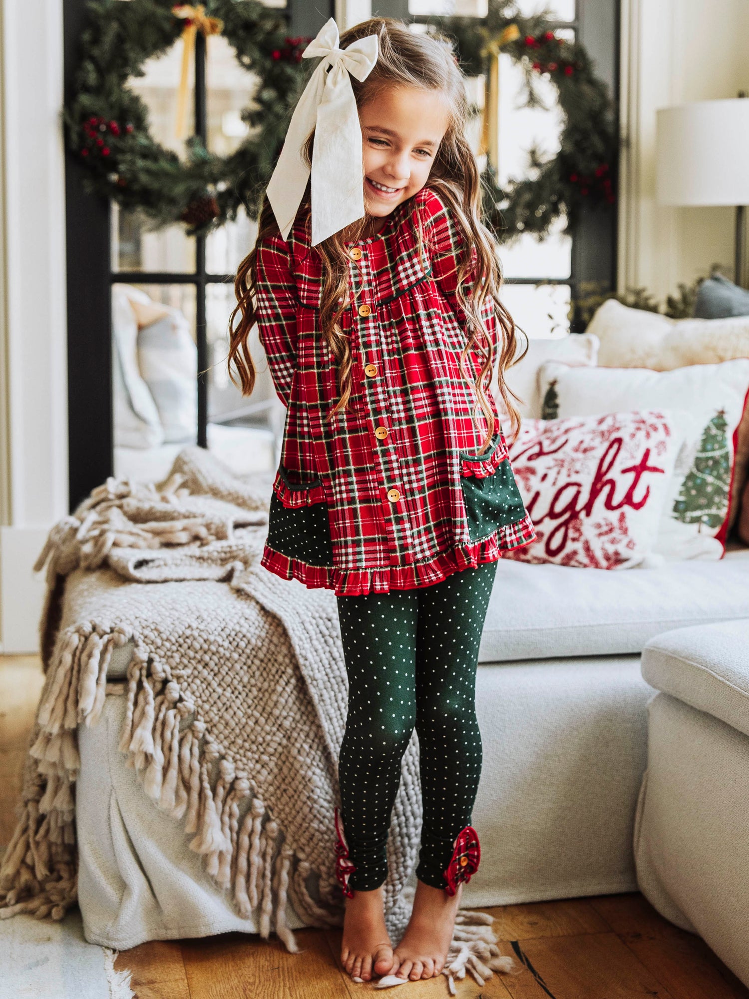 Girl wearing a two piece, Christmas play set. Top is a traditional plaid Christmas print and the bottoms are dark green polka dot. 