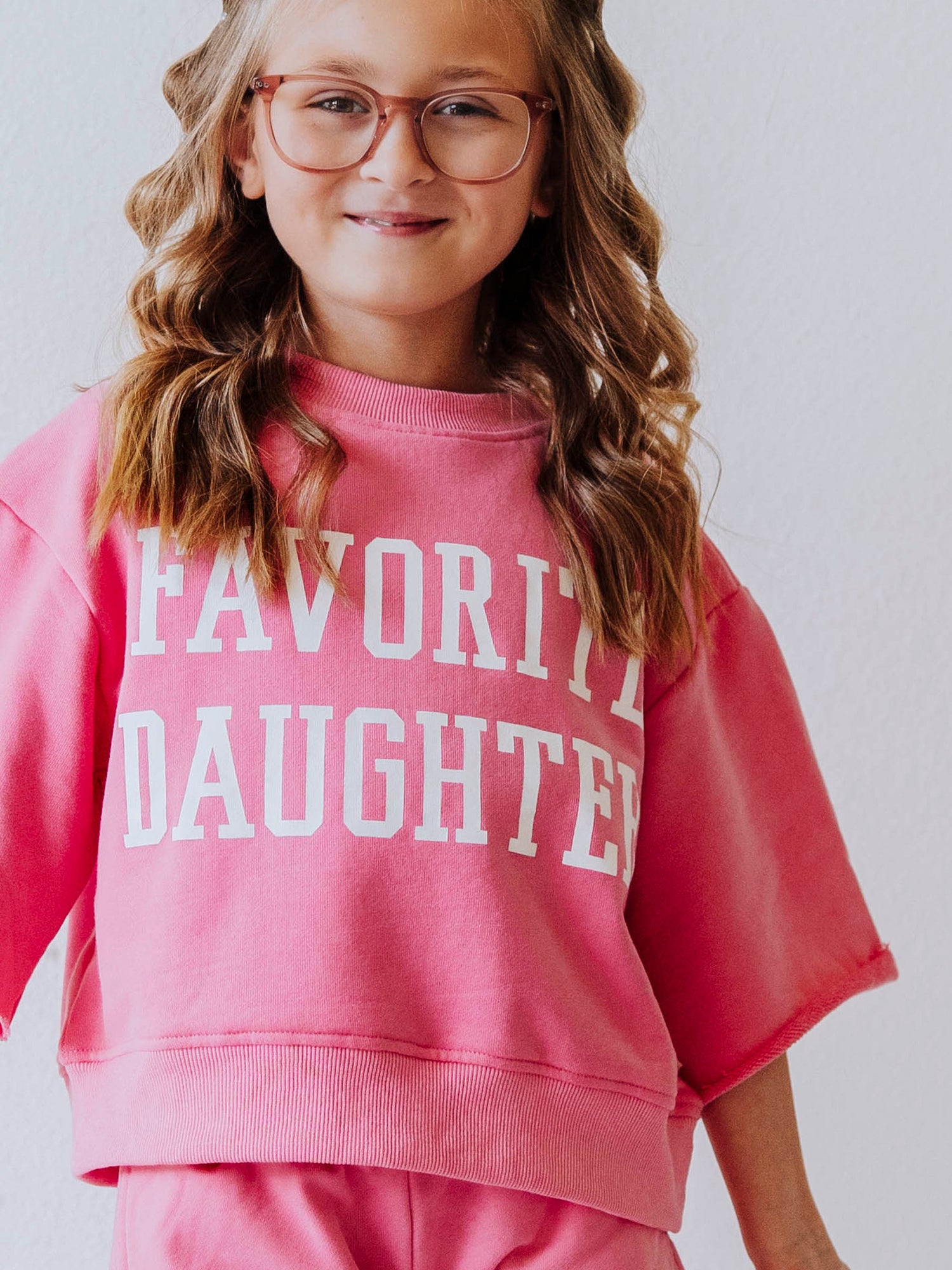 Close up of girl wearing a mock neck style shirt that is pink in color and has FAVORITE DAUGHTER written on the front in white vinyl lettering. 