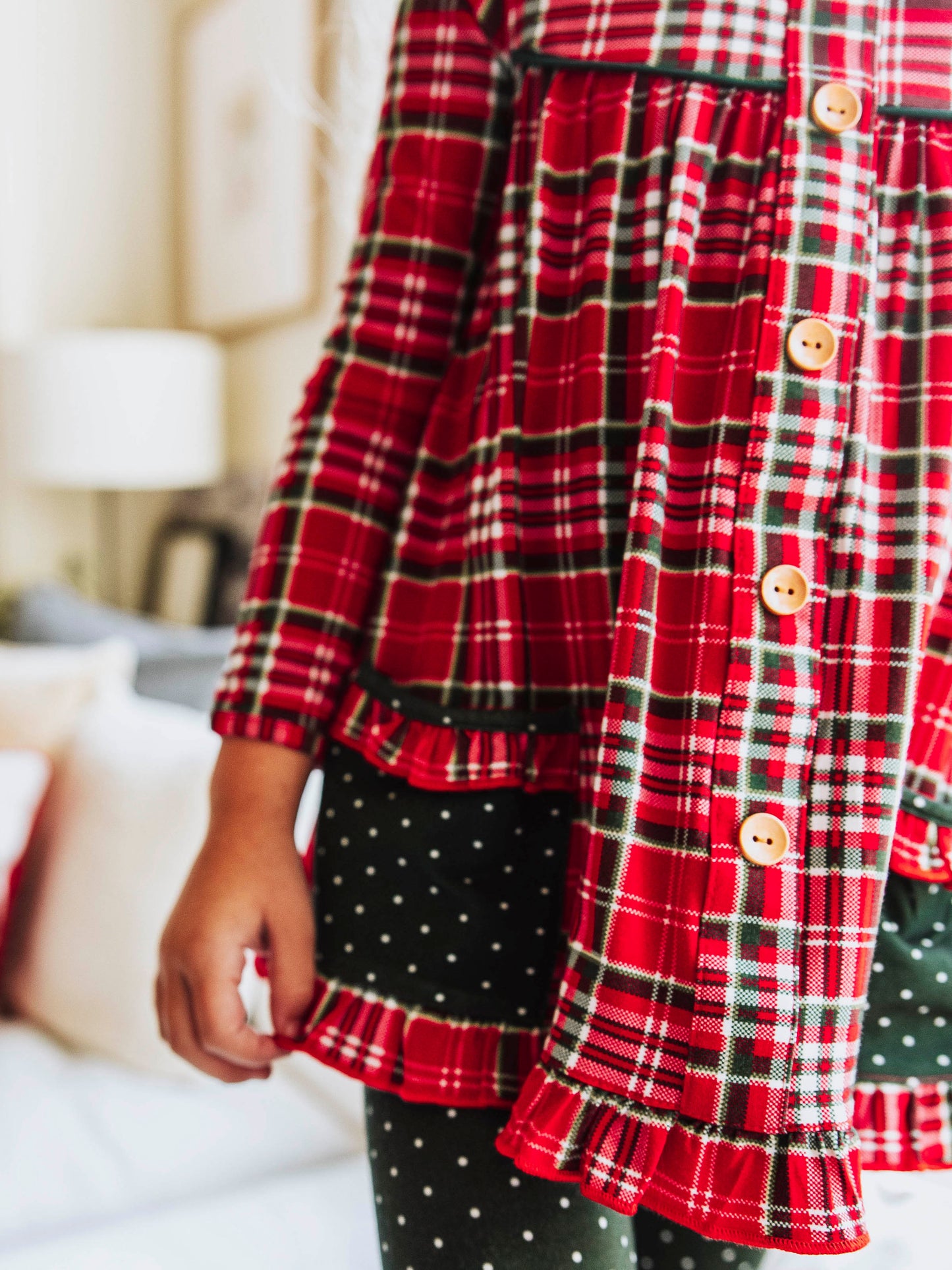 Girl wearing a two piece, Christmas play set. Top is a traditional plaid Christmas print and the bottoms are dark green polka dot. Up close shot of top with wooden buttons