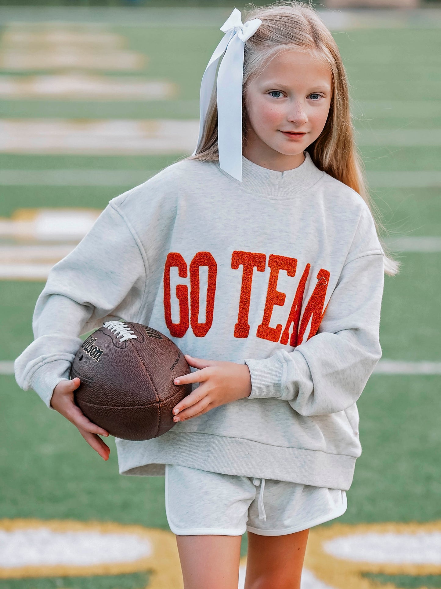 Girl outside wearing a heathered gray sweatshirt and shorts. The shirt says GO TEAM in burnt orange. Girl is holding a football