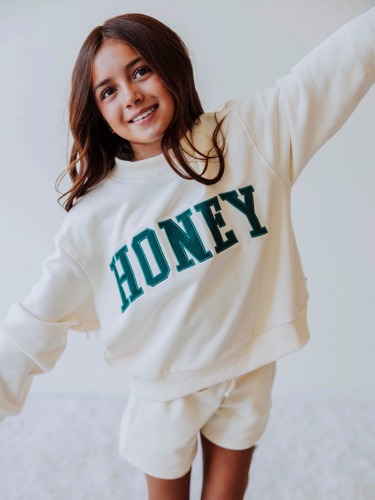 Girl model is wearing a two piece, ivory colored mock neck style set that has HONEY across the top in a darkish colored green. Bottoms she is wearing are the same ivory colored top. 
