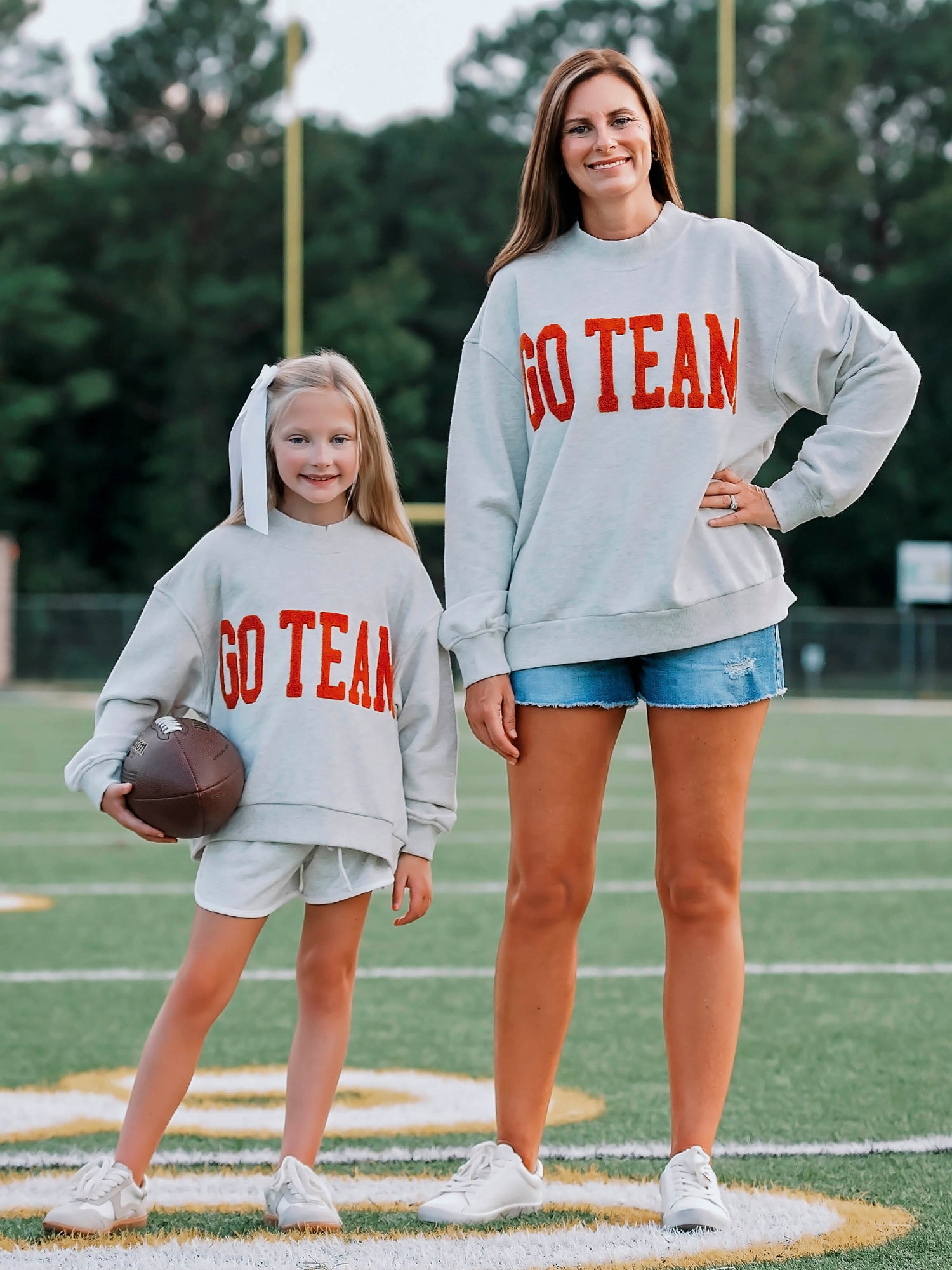 Women outside wearing a heathered gray sweater with GO TEAM on the front in burnt orange. She is also wearing denim jeans, A little girl is beside her and is wearing the same sweater and holding a football.