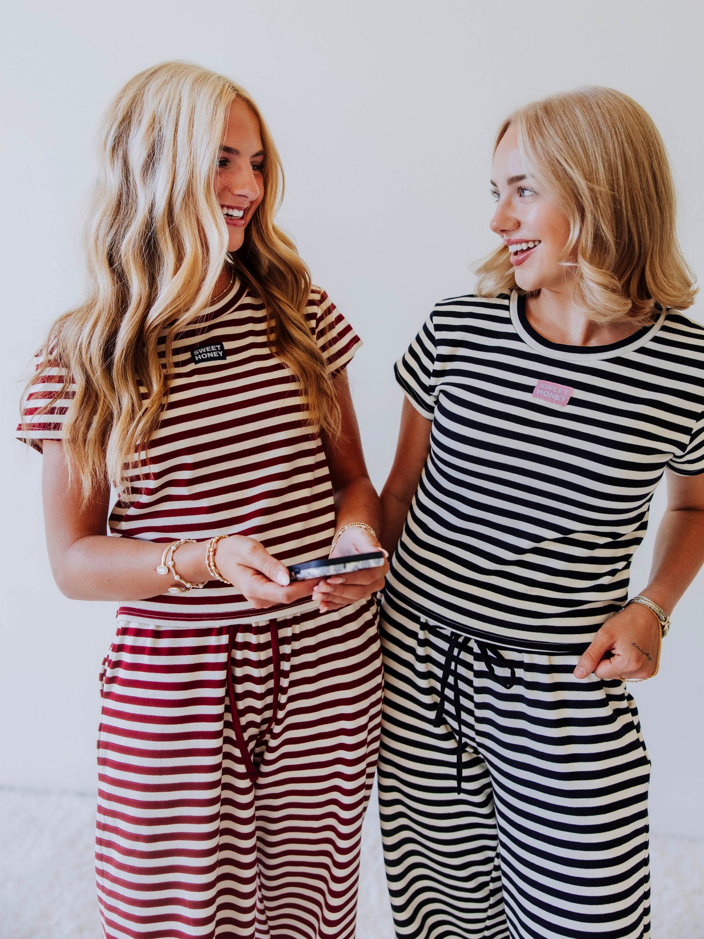 Two models both wearing pants sets in different colors. One is a crimson and ivory set and that model is holding a phone. Other model is in a navy and ivory colored pant set. 