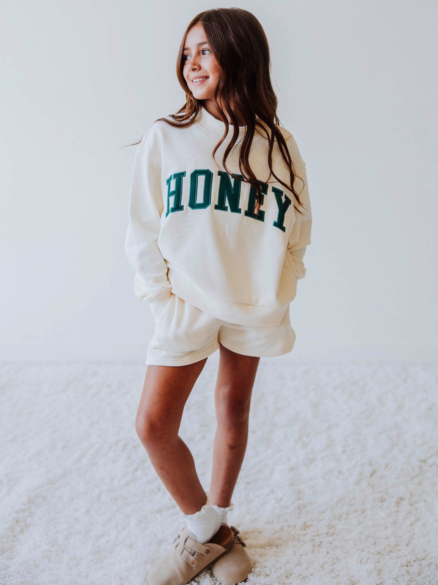 Girl model is wearing a two piece, ivory colored mock neck style set that has HONEY across the top in a darkish colored green. Bottoms she is wearing are the same ivory colored top. Model is wearing socks with clogs. 