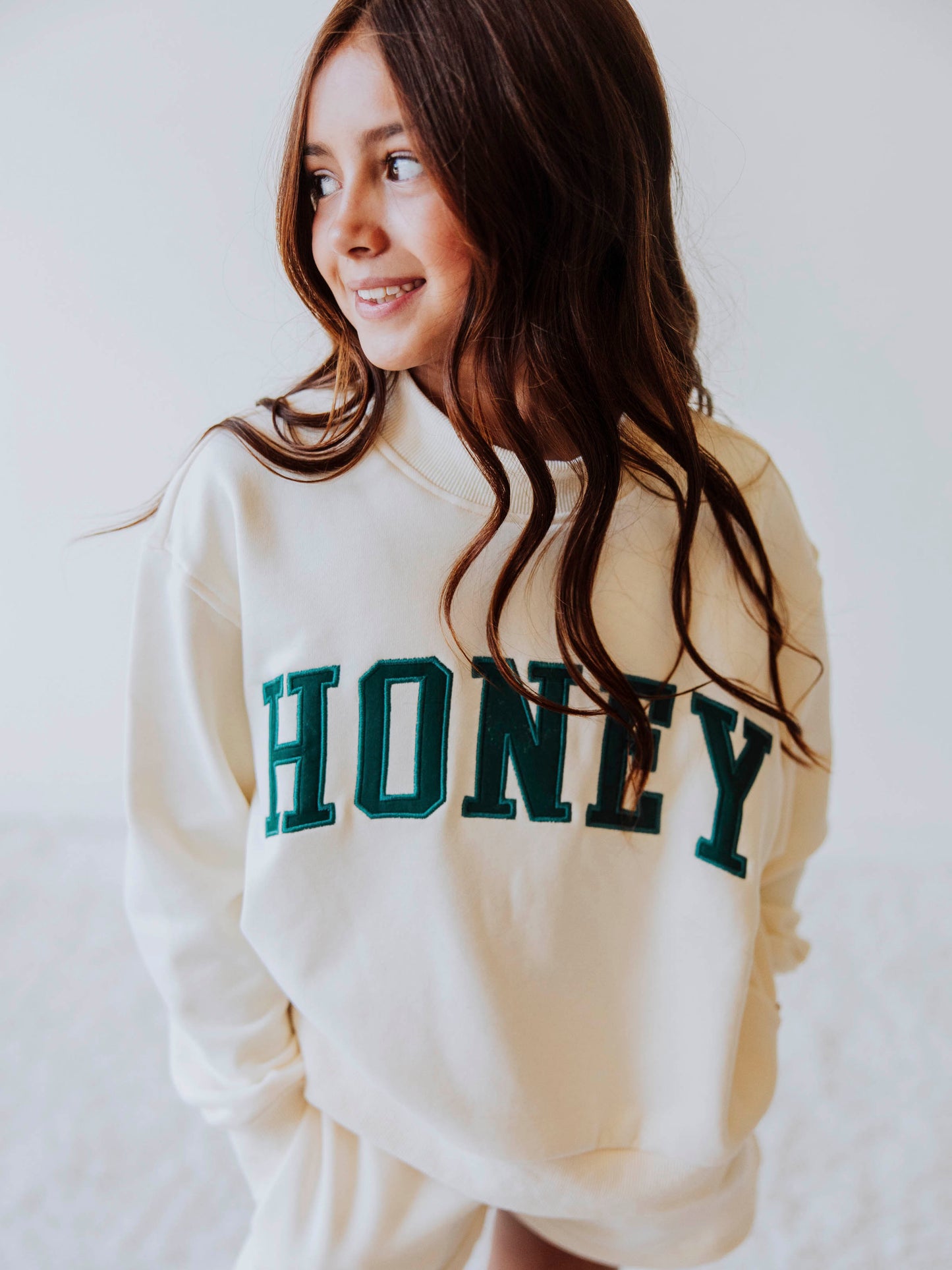 Girl model is wearing an ivory, mock neck style shirt that has HONEY on the front in dark greenish letters. 