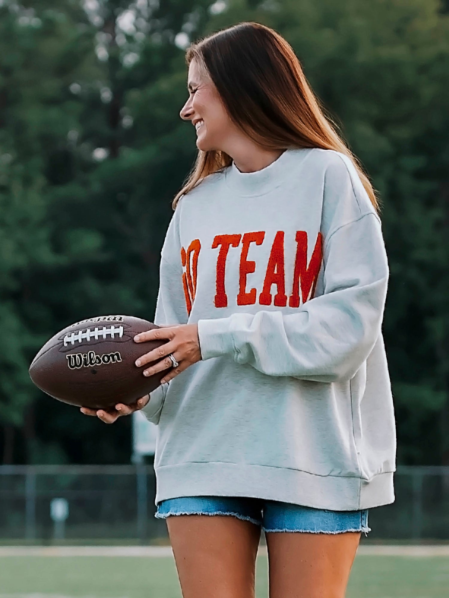 Women outside wearing a heathered gray sweater with GO TEAM on the front in burnt orange. She is also wearing denim jeans and holding a football