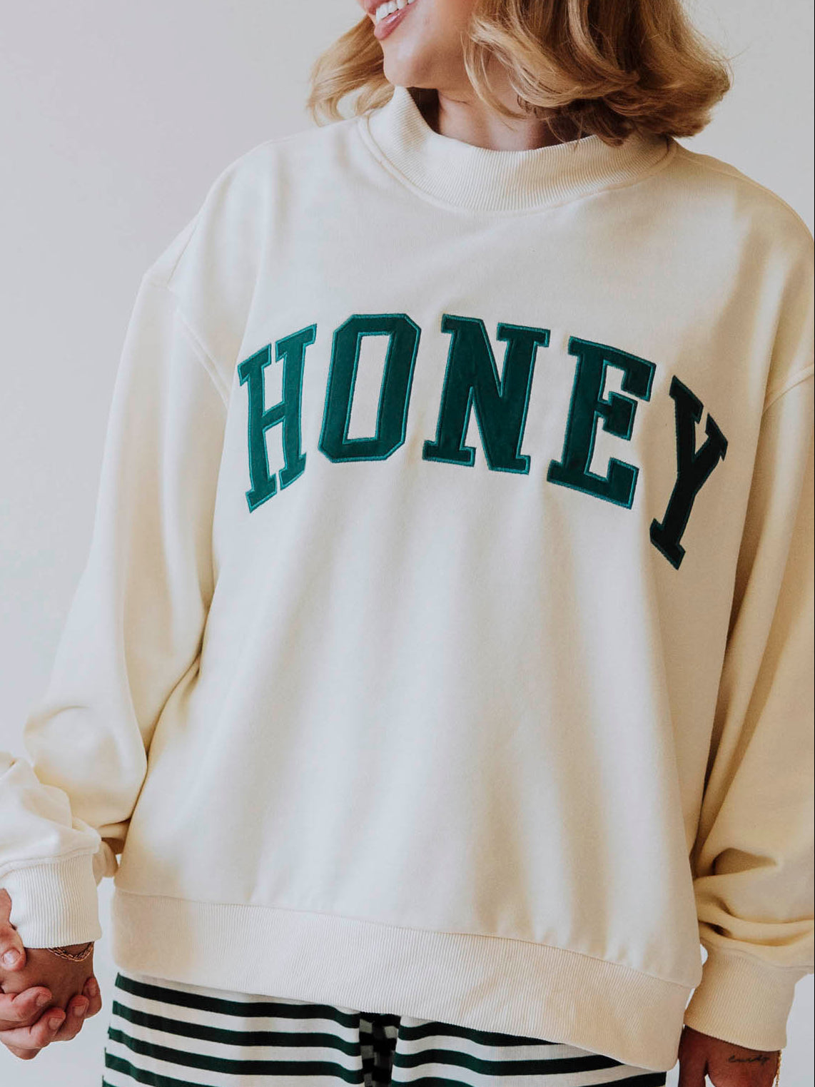 Girl wearing a cream mock neck sweatshirt that says HONEY in green.