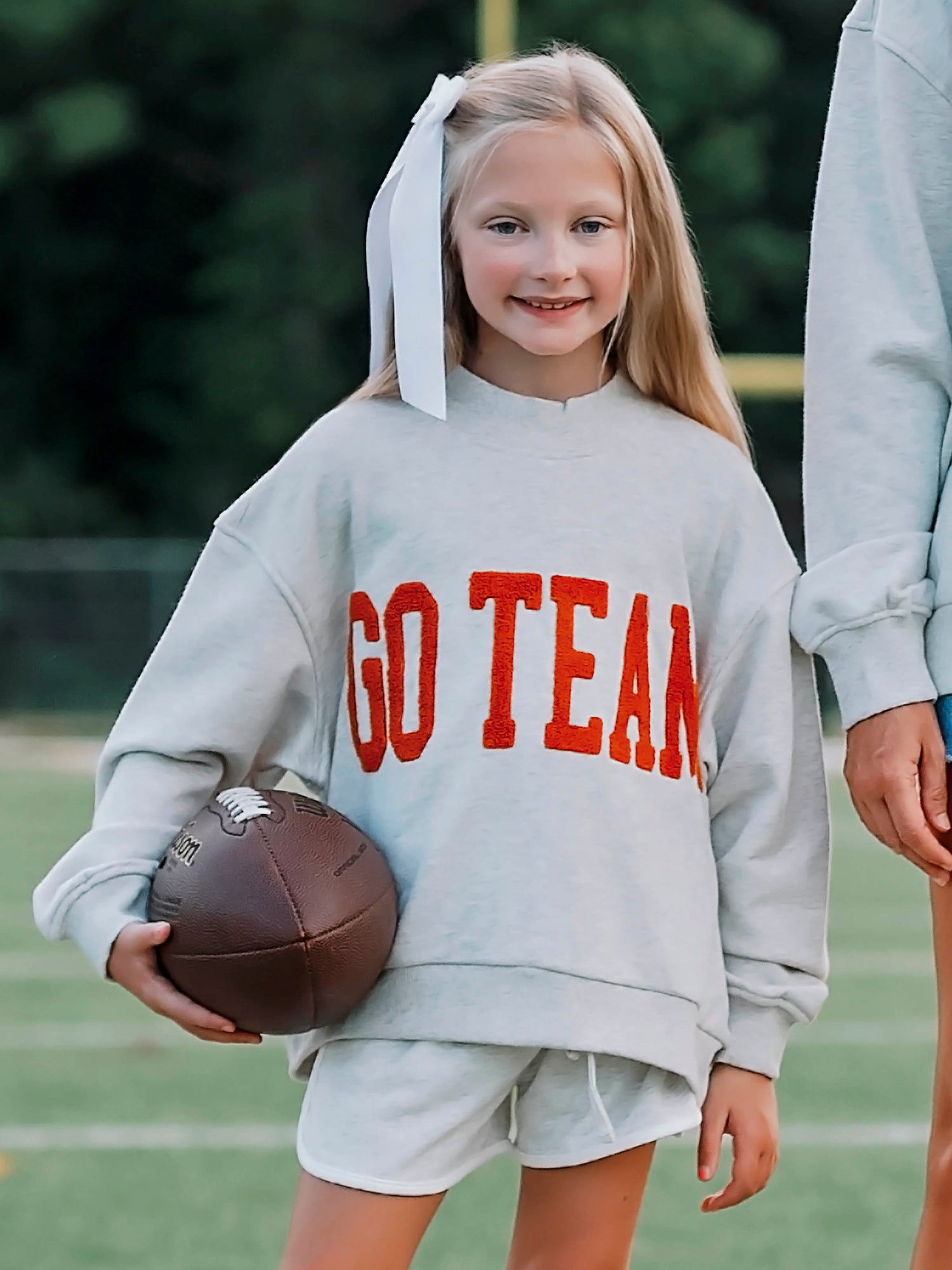 Girl outside wearing a heathered gray sweatshirt and shorts. The shirt says GO TEAM in burnt orange. Girl is holding a football