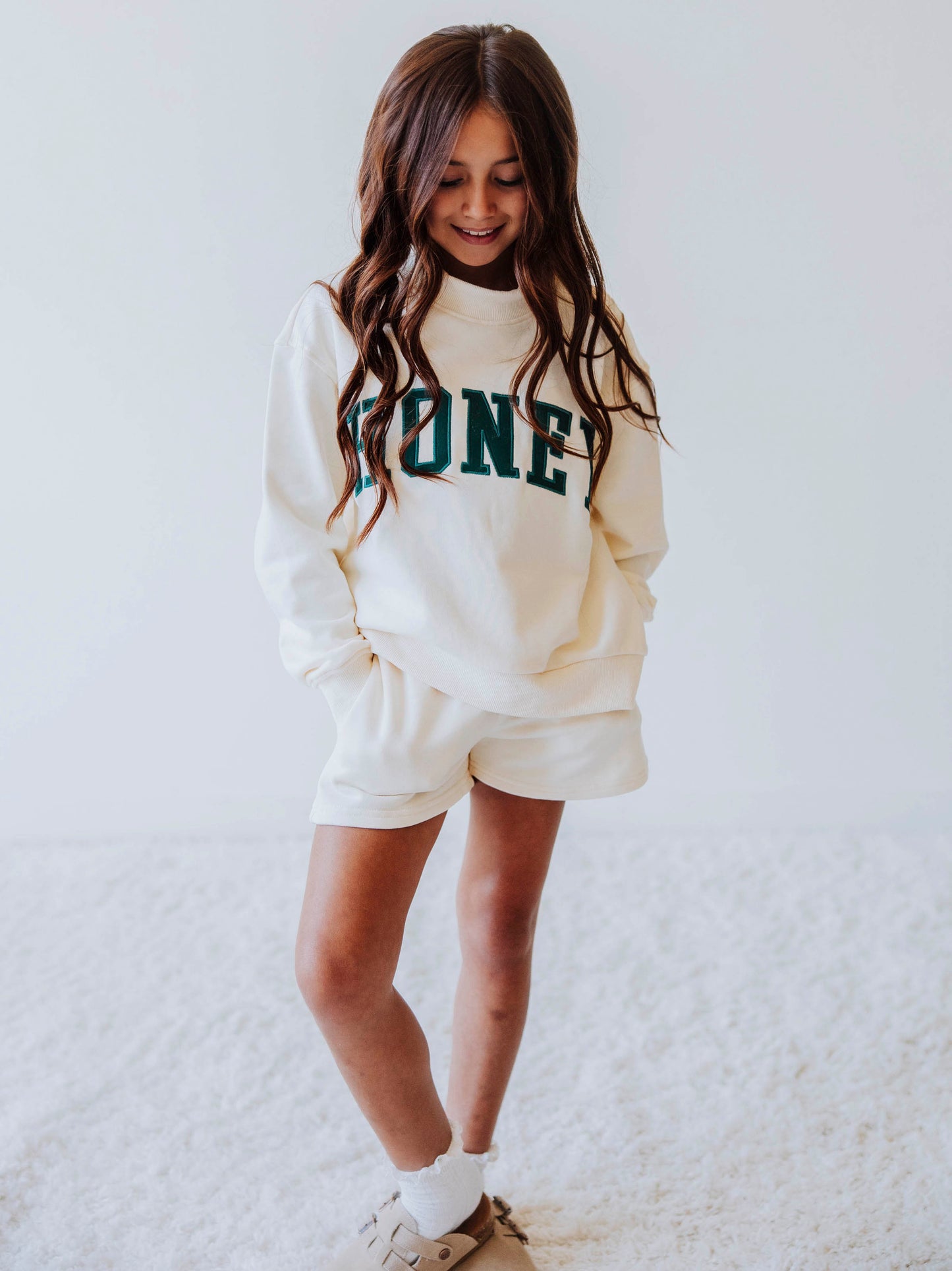 Girl model is wearing a two piece, ivory colored mock neck style set that has HONEY across the top in a darkish colored green. Bottoms she is wearing are the same ivory colored top. Model is wearing socks with clogs. 