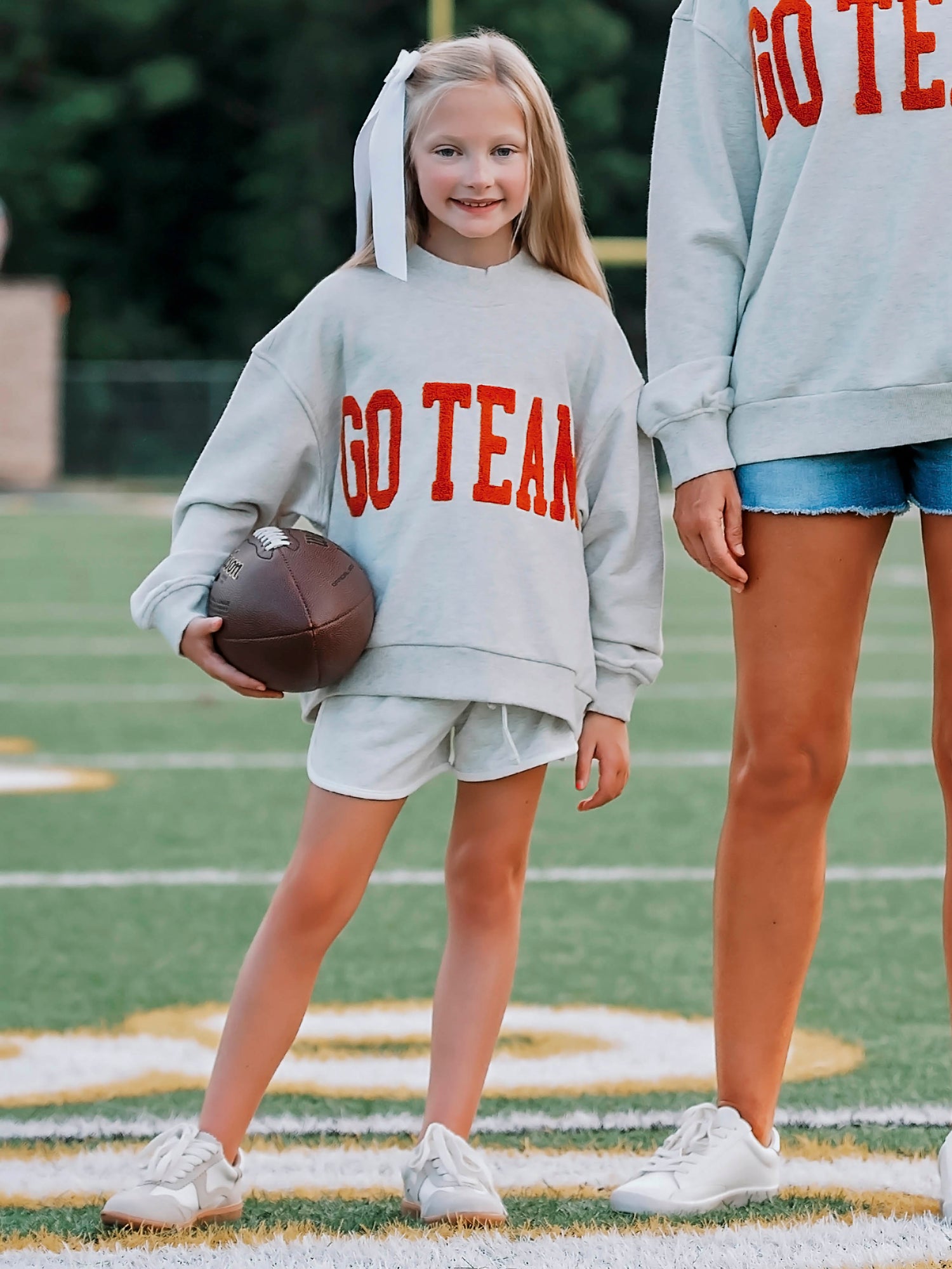 Girl outside wearing a heathered gray sweatshirt and shorts. The shirt says GO TEAM in burnt orange. Girl is holding a football and standing next to an adult. 