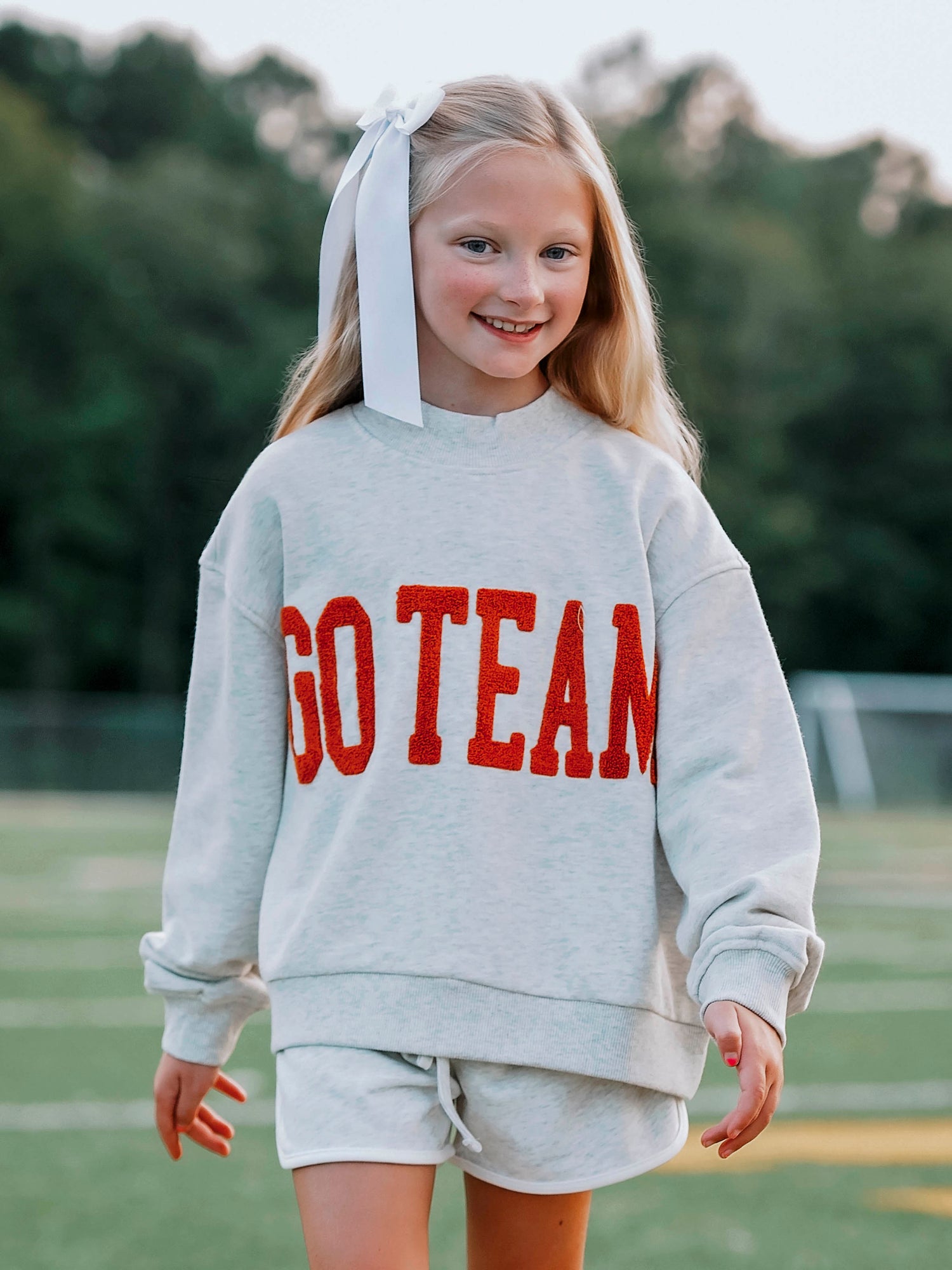 Girl outside wearing a heathered gray sweatshirt and shorts. The shirt says GO TEAM in burnt orange. 