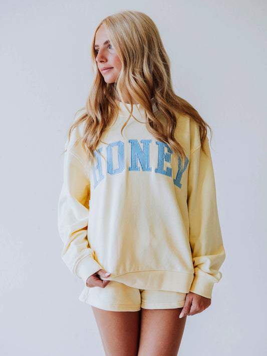 Girl wearing a soft, yellow two piece set. Top says HONEY on the front. The HONEY has blue lines going through each letter. Shorts are same yellow as top. 