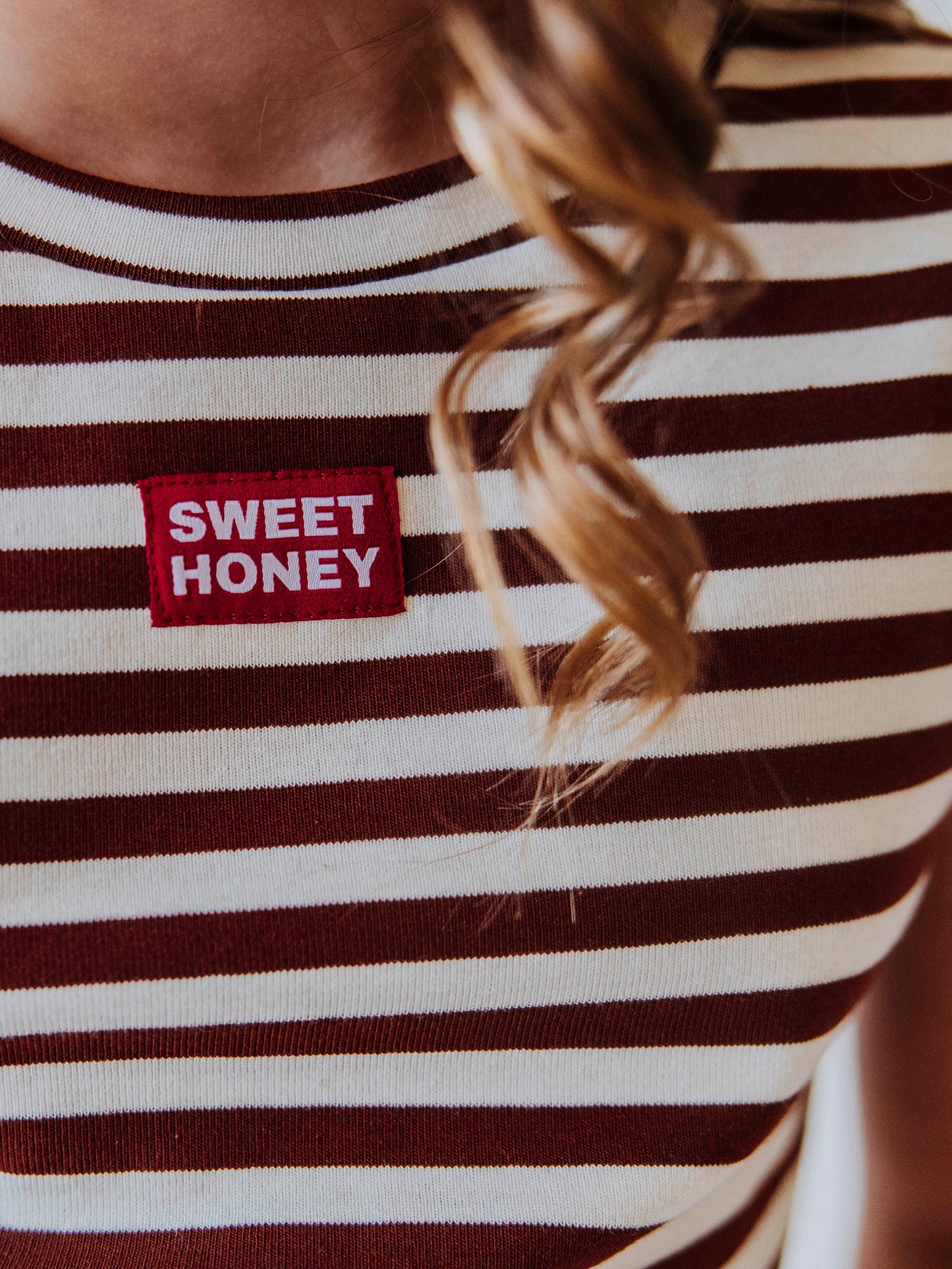 Up close shot of a red and white SweetHoney tag on chest. Color of shirt is blrown and ivory striped. 
