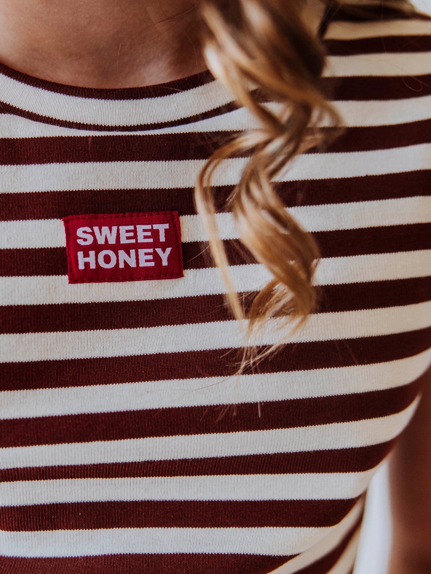 Up close shot of a red and white SweetHoney tag on chest. Color of shirt is blrown and ivory striped. 

