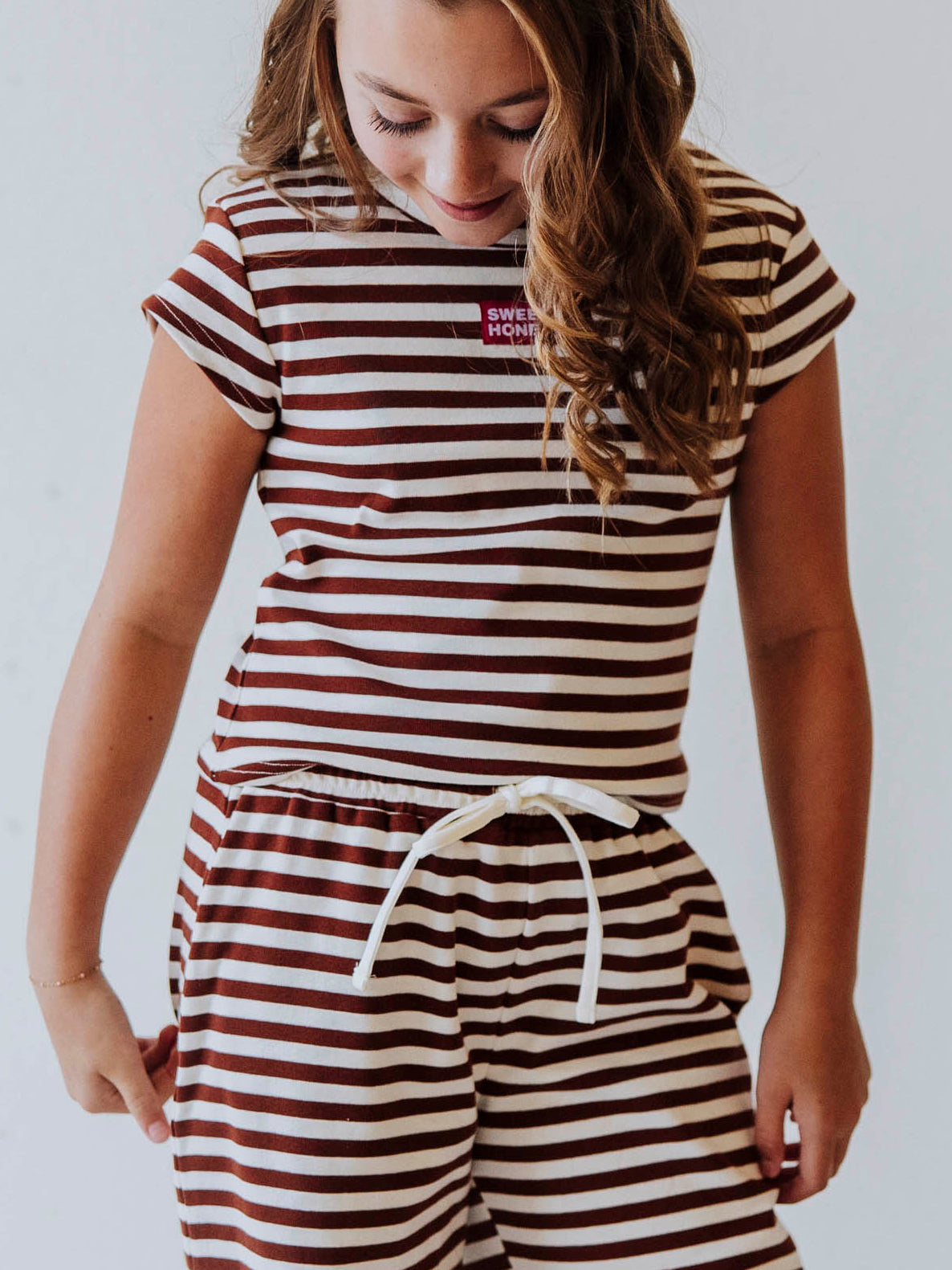 Girl wearing a two-piece brown and ivory striped pant set with clogs. Shirt has a SweetHoney tag on chest that is red and white. 