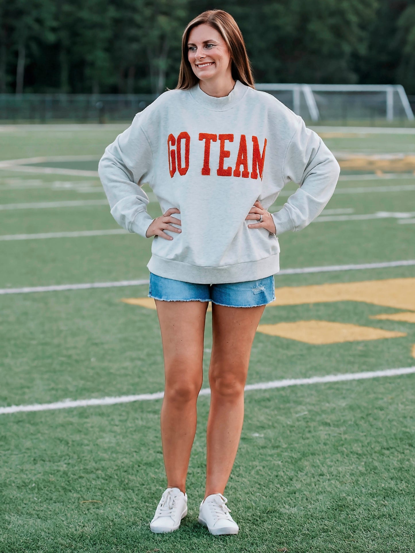 Women outside wearing a heathered gray sweater with GO TEAM on the front in burnt orange. She is also wearing denim jeans. 