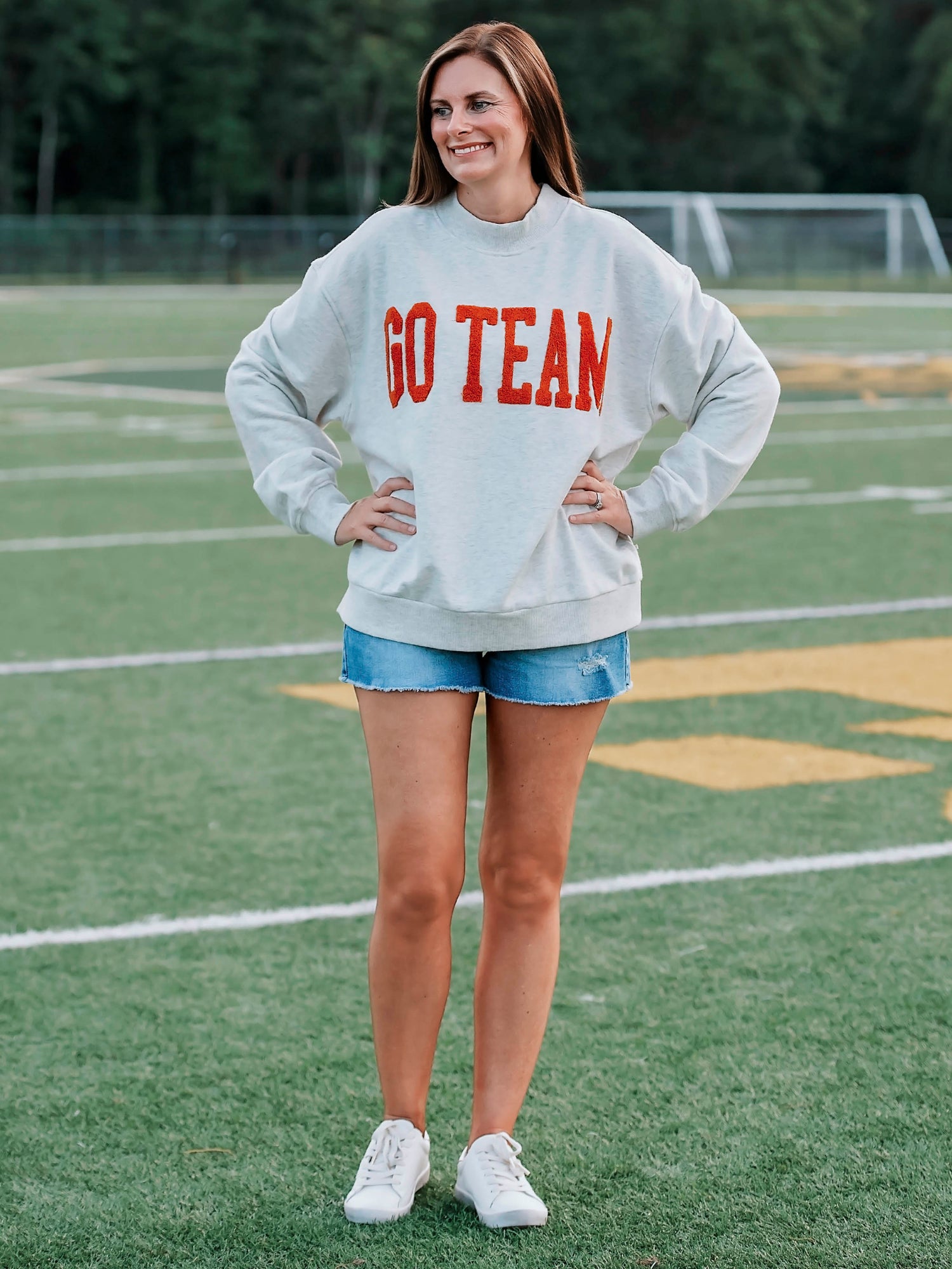 Women outside wearing a heathered gray sweater with GO TEAM on the front in burnt orange. She is also wearing denim jeans. 