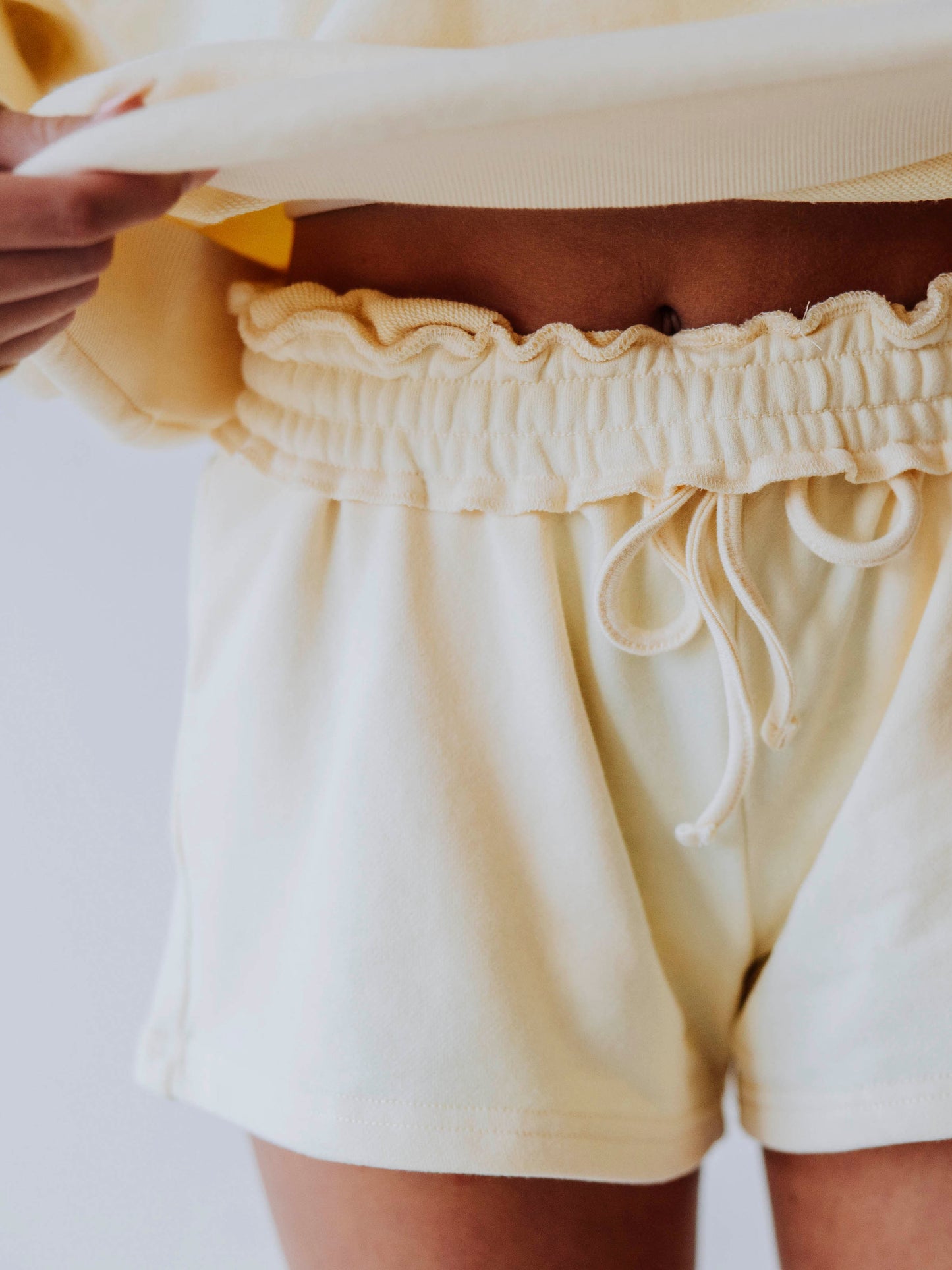 Photo shows a soft yellow pair of shorts on model. Waistband is rolled one time and drawstring is showing as well. Girl is holding up shirt to show this. 