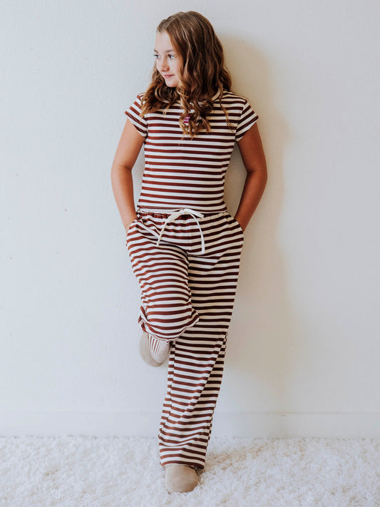 Girl wearing a two-piece brown and ivory striped pant set with clogs. Foot is propped up against the wall. 