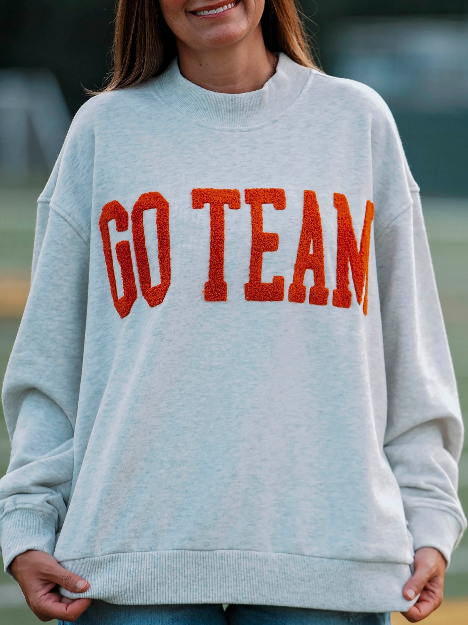 Women outside wearing a heathered gray sweater with GO TEAM on the front in burnt orange. 