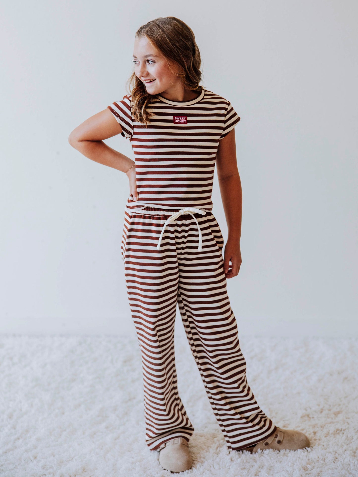 Girl wearing a two-piece brown and ivory striped pant set with clogs. There is a SweetHoney tag on chest of shirt. 