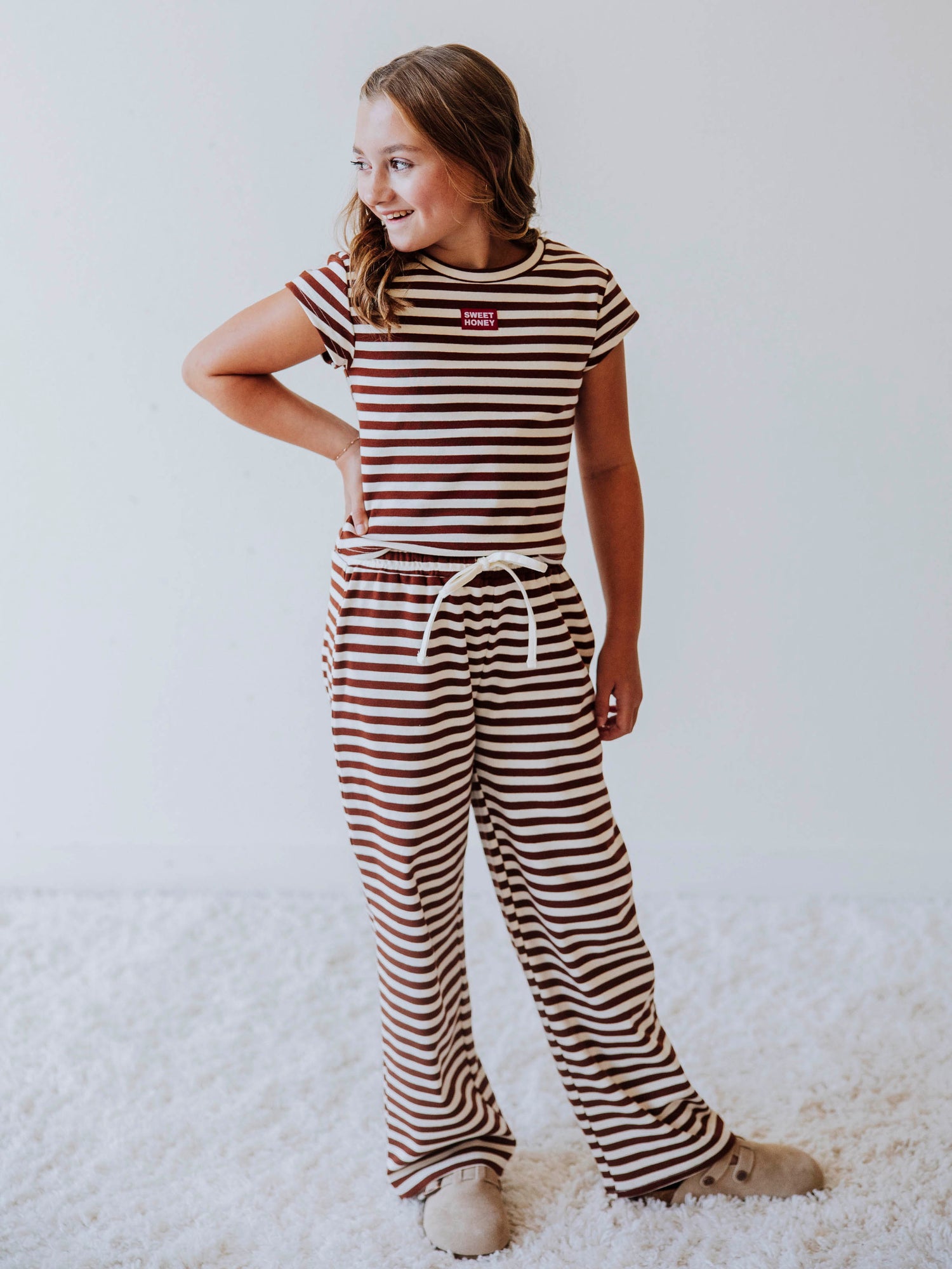 Girl wearing a two-piece brown and ivory striped pant set with clogs. There is a SweetHoney tag on chest of shirt. 
