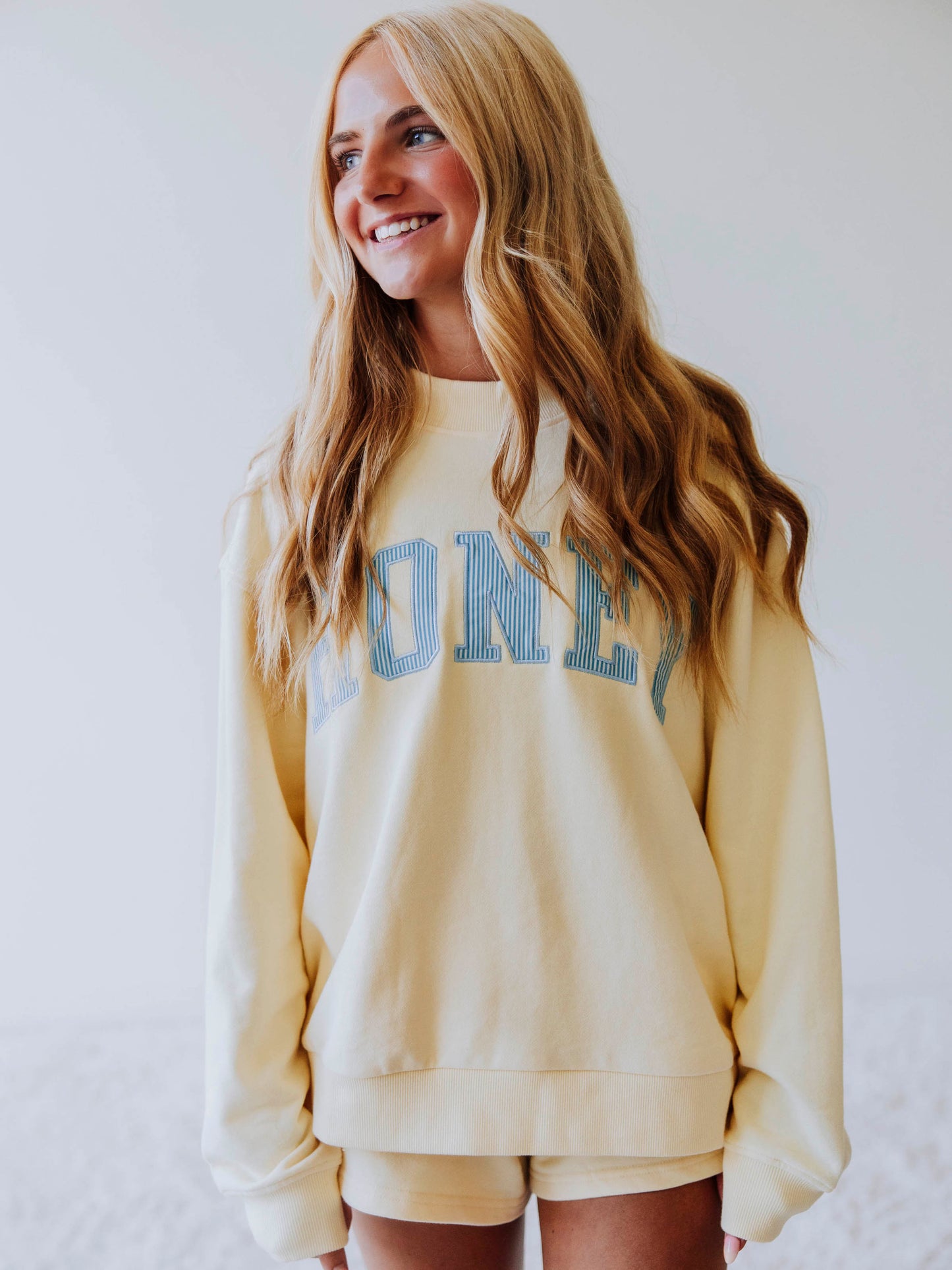 Girl wearing a soft, yellow two piece set. Top says HONEY on the front. The HONEY has blue lines going through each letter. Shorts are same yellow as top. 